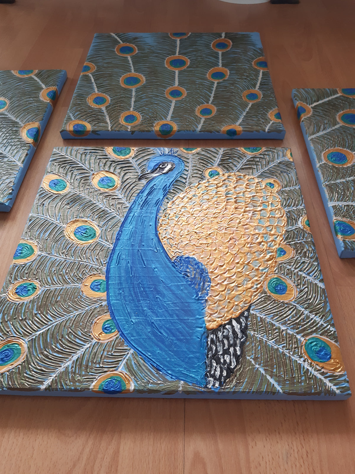 Vierteiliges handgefertigtes Leinwandbild-Set mit stilisiertem Pfauenmotiv in Blau, Türkis und Gold. Strukturierte Acrylmalerei auf Leinwand als einzigartiges Wandkunst-Unikat von Solas Artwork.