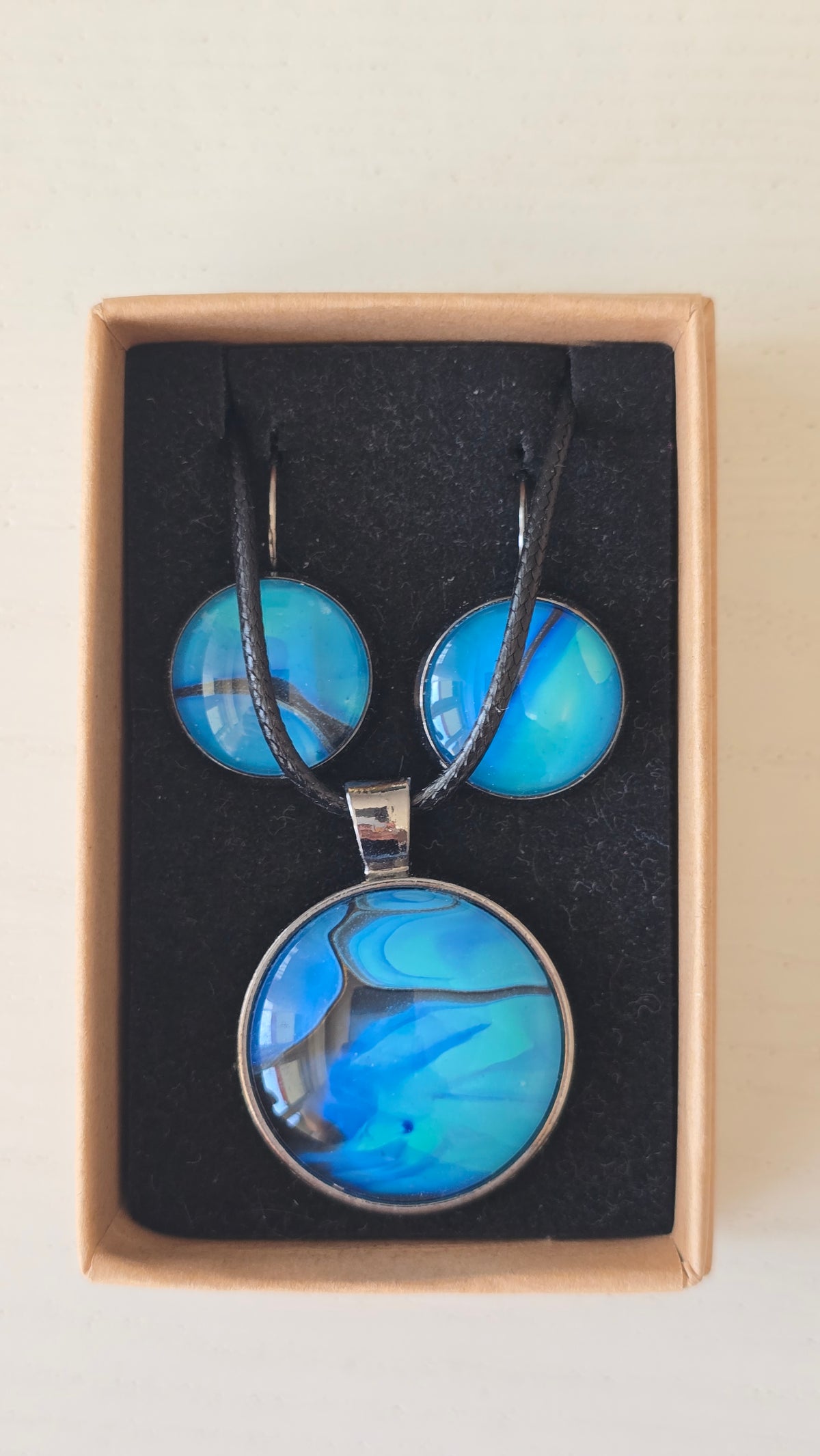 Handgefertigtes Schmuckset Solas Laguna mit rundem Anhänger und passenden Ohrringen in Blau- und Türkistönen mit fliessendem Marmormuster unter Glas.