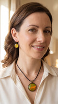 Frau trägt rundes Cabochon Schmuckset mit grossem Kettenanhänger und zwei passenden Ohrringen in Grün, Orange und Gold mit fliessenden, organischen Mustern, bronzefarbenen Fassungen und schwarzem Band.