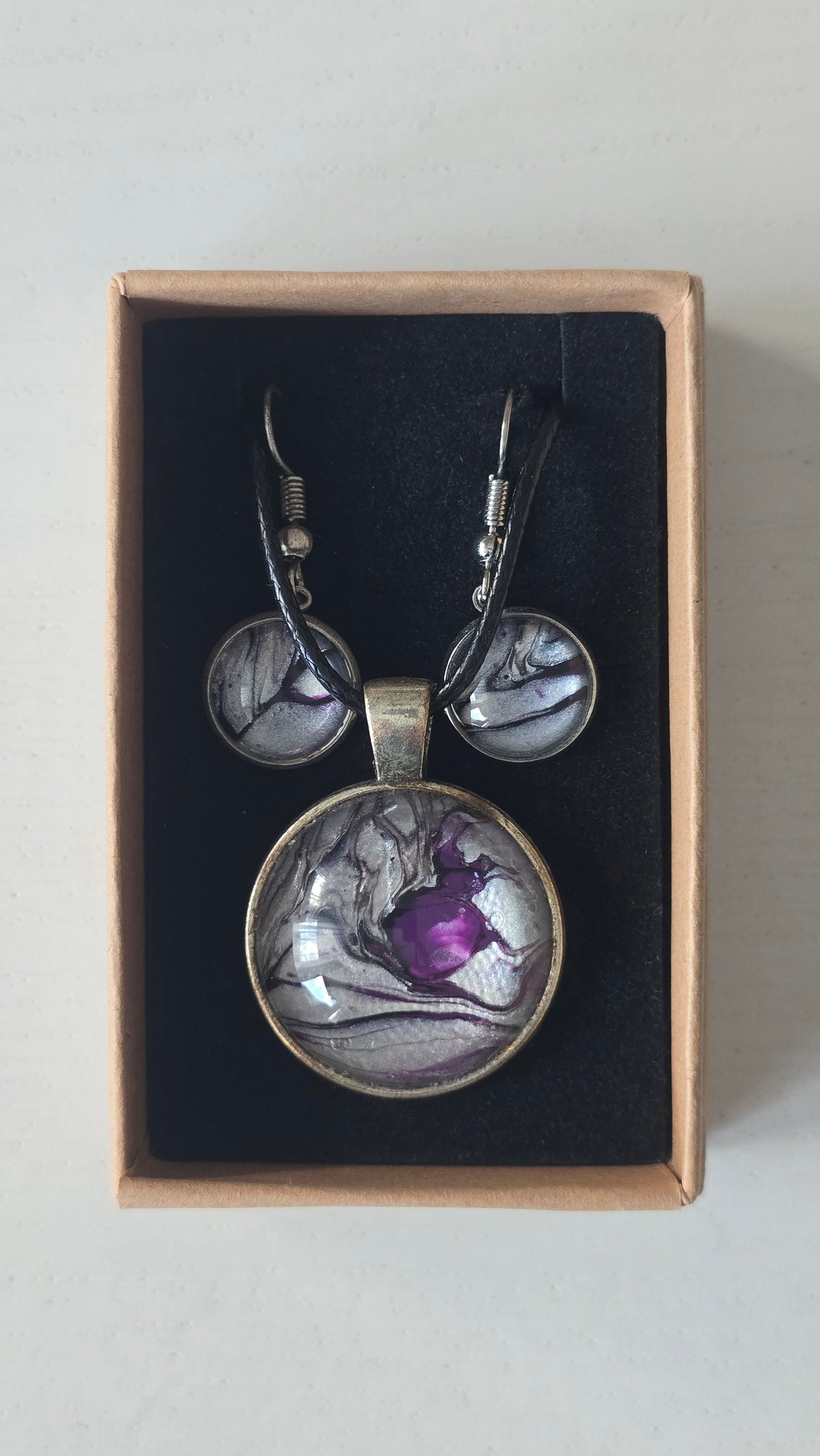 Cabochon-Schmuckset mit rundem Anhänger und zwei Ohrringen in schimmerndem Silber und Grau mit dunklen marmorierten Adern und violettem Farbkern.