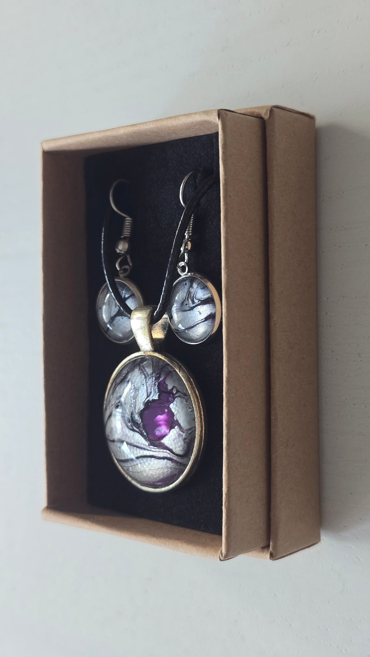 Cabochon-Schmuckset mit rundem Anhänger und zwei Ohrringen in schimmerndem Silber und Grau mit dunklen marmorierten Adern und violettem Farbkern.