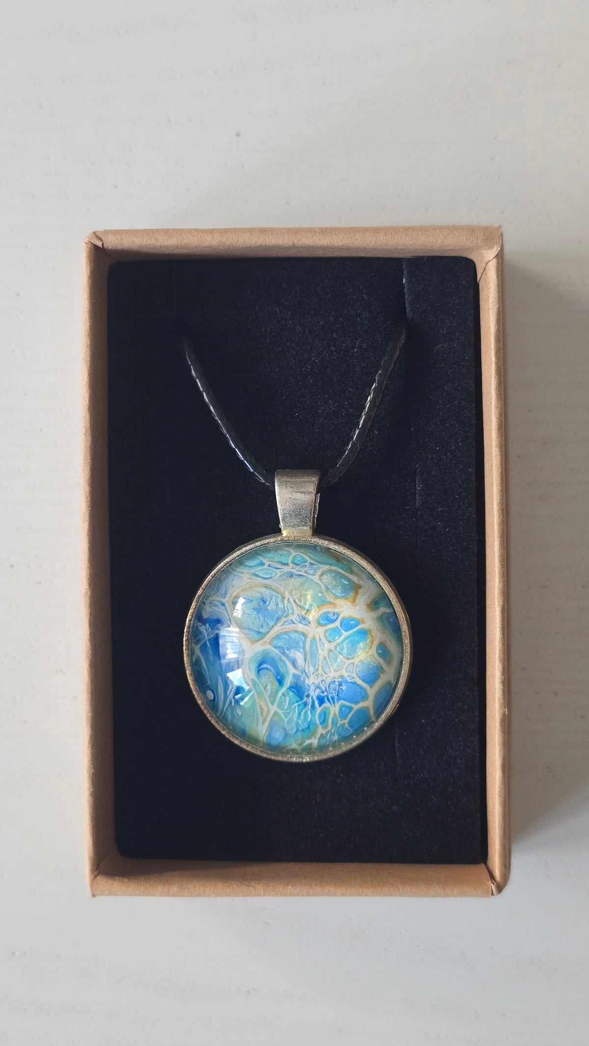 Runder Cabochon-Anhänger an schwarzem Band mit filigranem, netzartigem Muster in Aqua-, Türkis-, Hellblau- und Cremetönen.