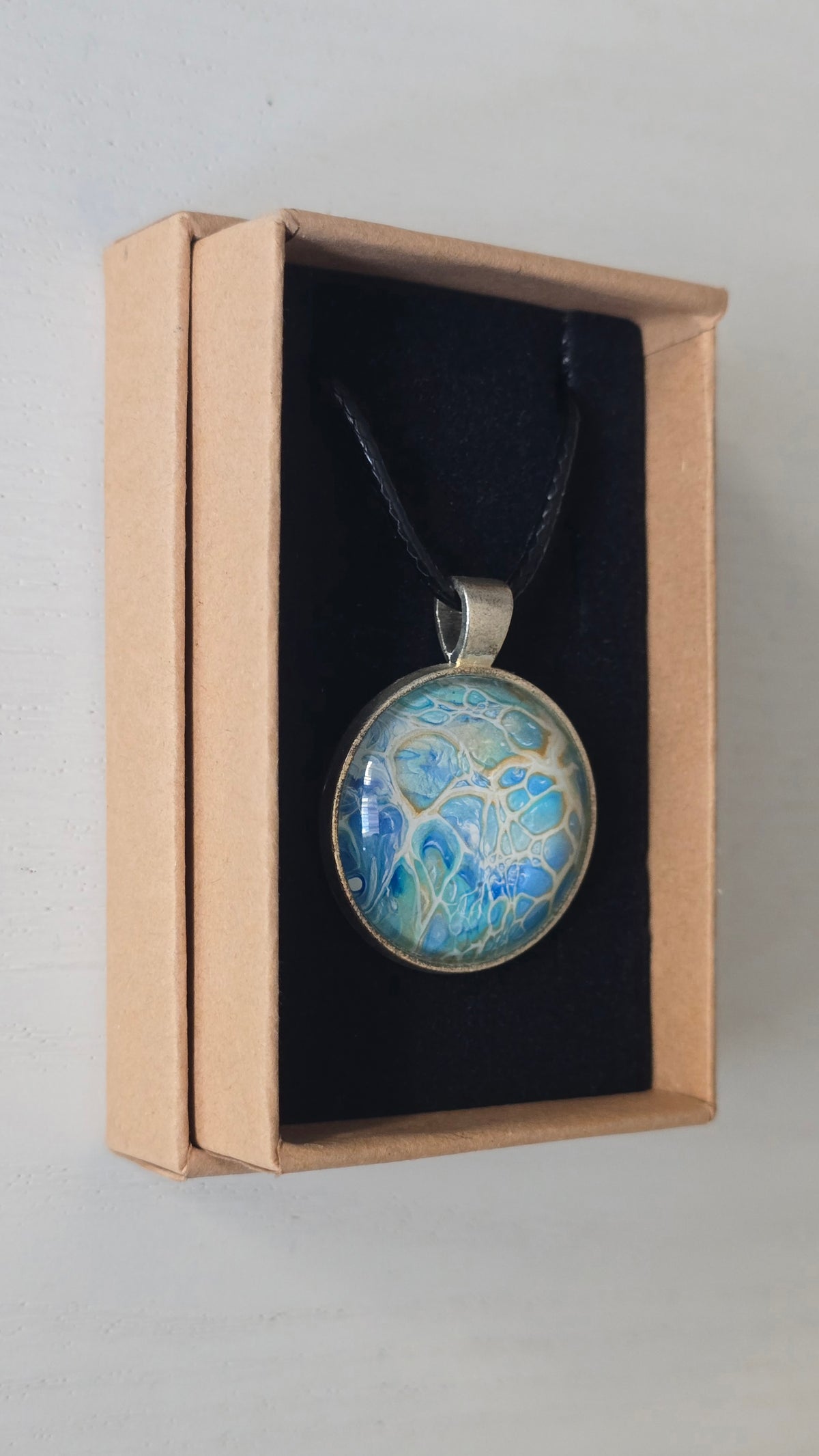 Runder Cabochon-Anhänger an schwarzem Band mit filigranem, netzartigem Muster in Aqua-, Türkis-, Hellblau- und Cremetönen.