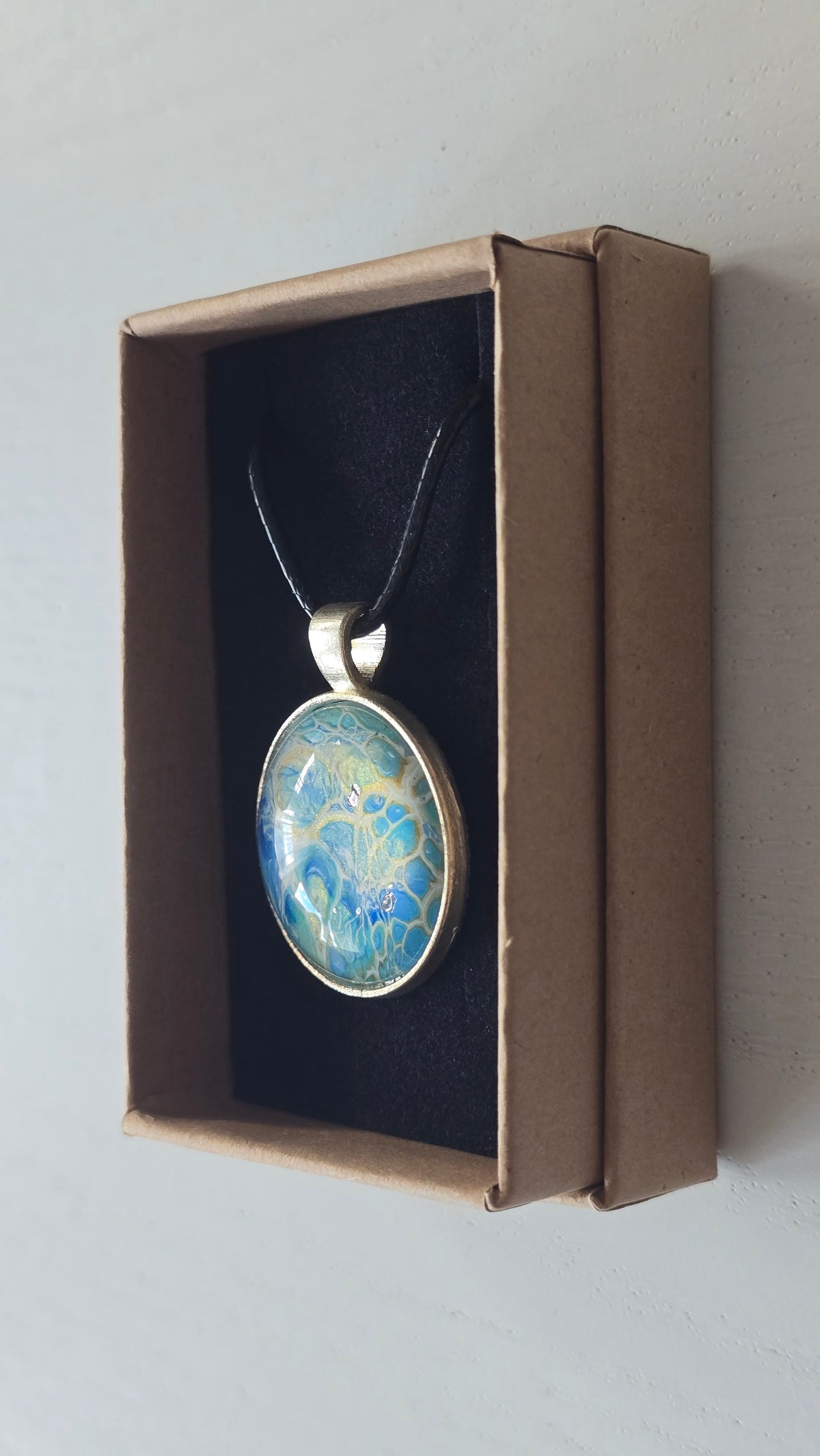 Runder Cabochon-Anhänger an schwarzem Band mit filigranem, netzartigem Muster in Aqua-, Türkis-, Hellblau- und Cremetönen.