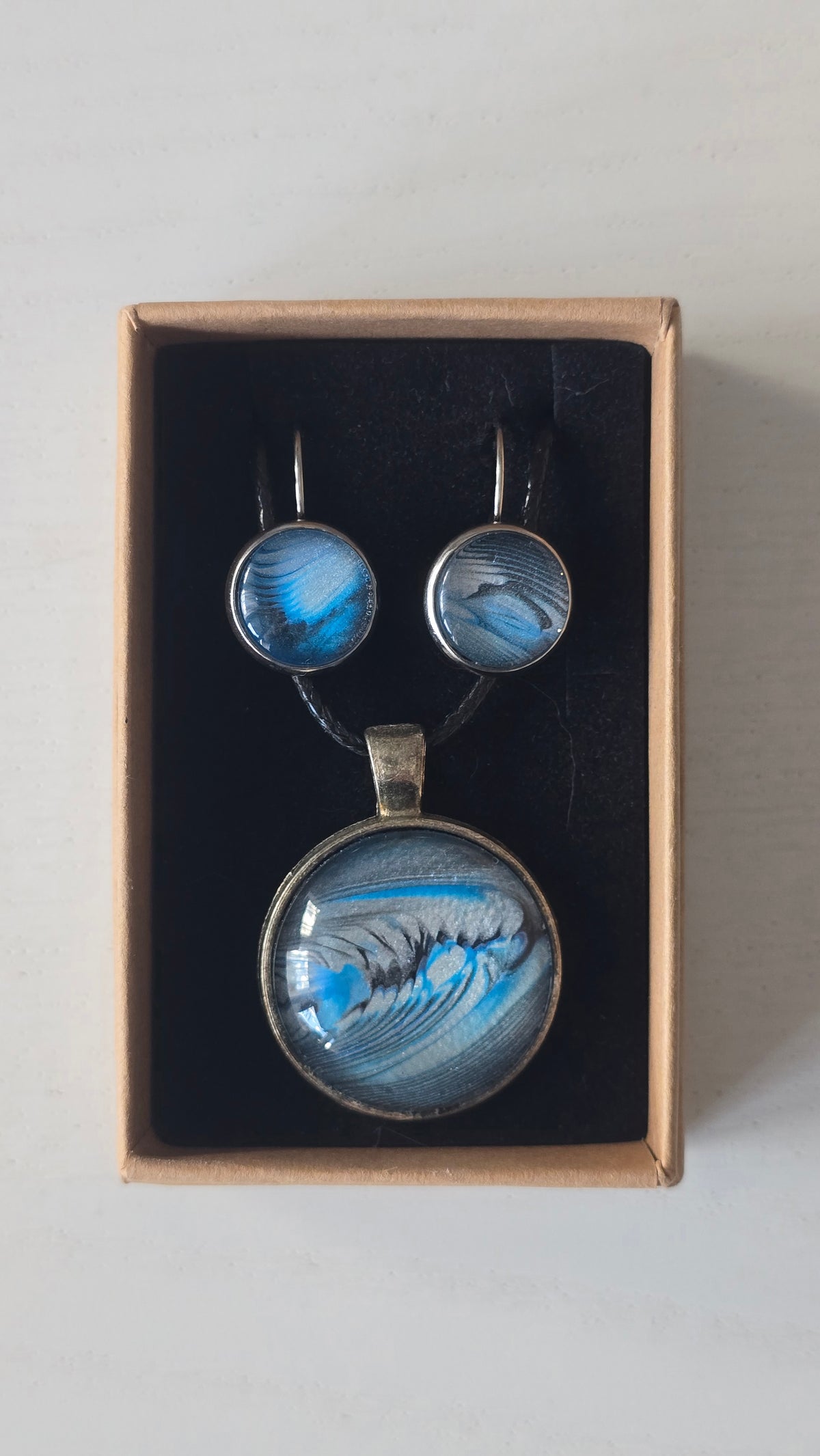 Rundes Cabochon-Schmuckset mit Anhänger und Ohrringen an schwarzem Band, mit fliessendem abstraktem Muster in Cyan, Blau, Grau und Schwarz.