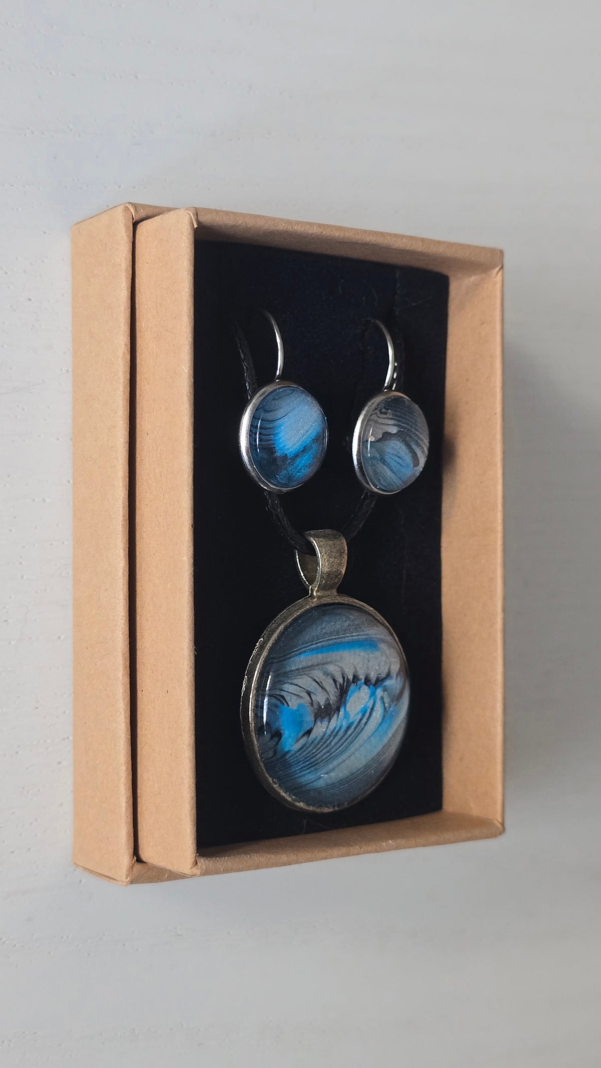 Rundes Cabochon-Schmuckset mit Anhänger und Ohrringen an schwarzem Band, mit fliessendem abstraktem Muster in Cyan, Blau, Grau und Schwarz.