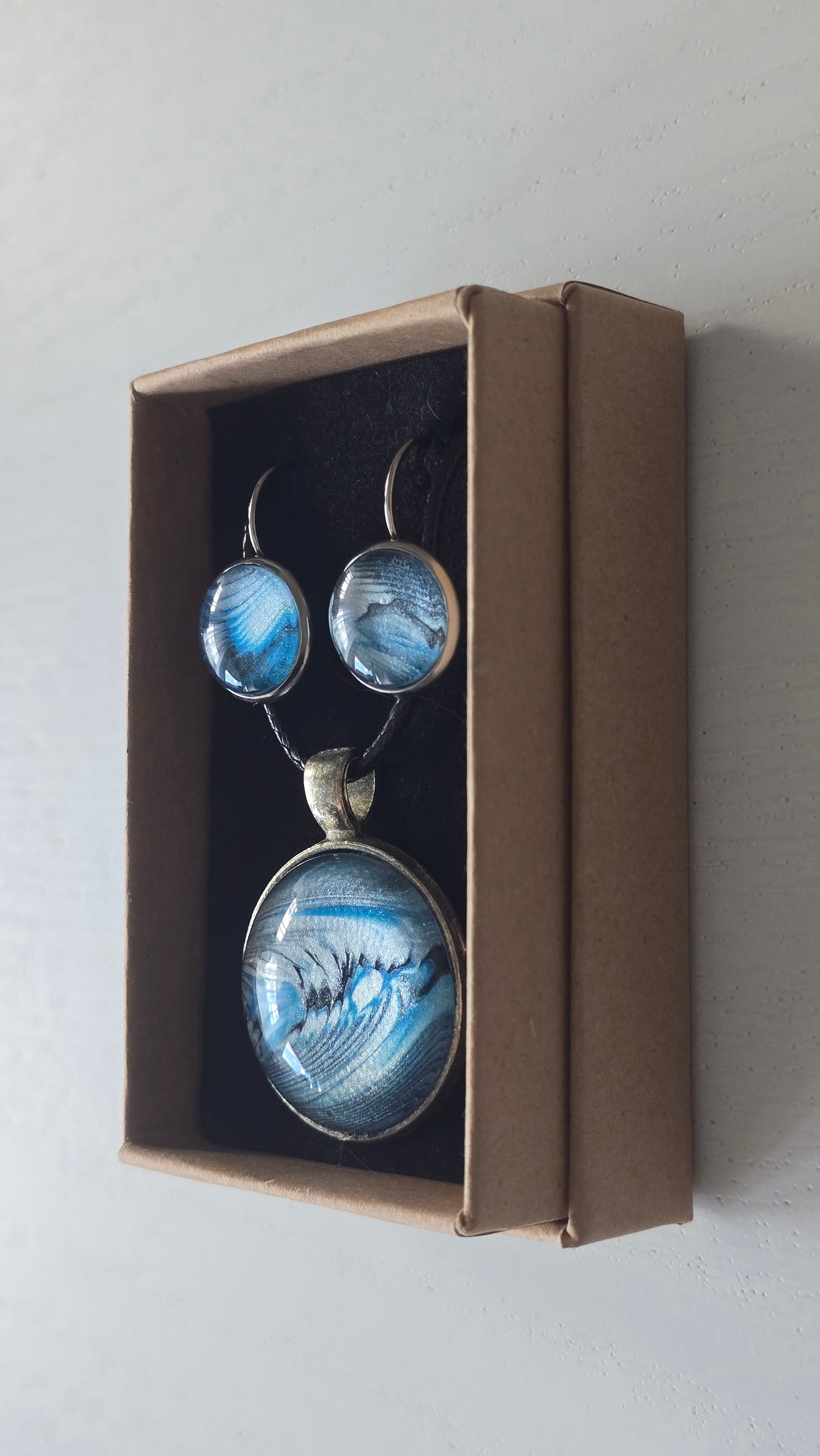 Rundes Cabochon-Schmuckset mit Anhänger und Ohrringen an schwarzem Band, mit fliessendem abstraktem Muster in Cyan, Blau, Grau und Schwarz.