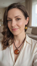 Frau trägt Cabochon-Schmuckset mit rundem Anhänger und zwei Ohrringen mit organischem Muster in Rostrot, Orange und leuchtendem Grün, kupferfarbenen Fassungen und schwarzem Textilband.