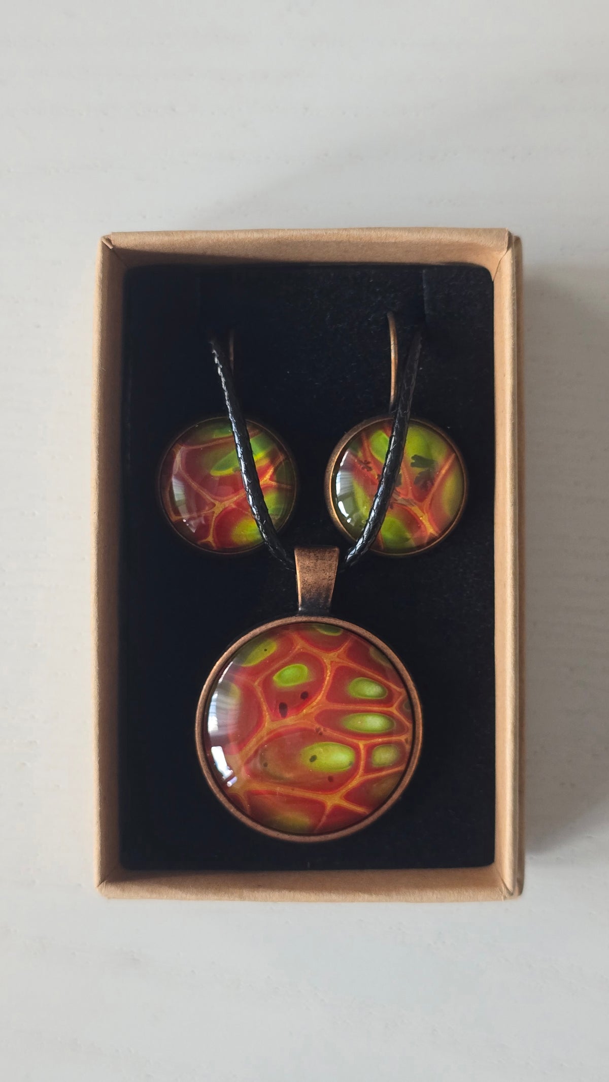 Cabochon-Schmuckset mit rundem Anhänger und zwei Ohrringen mit organischem Muster in Rostrot, Orange und leuchtendem Grün, kupferfarbenen Fassungen und schwarzem Textilband.