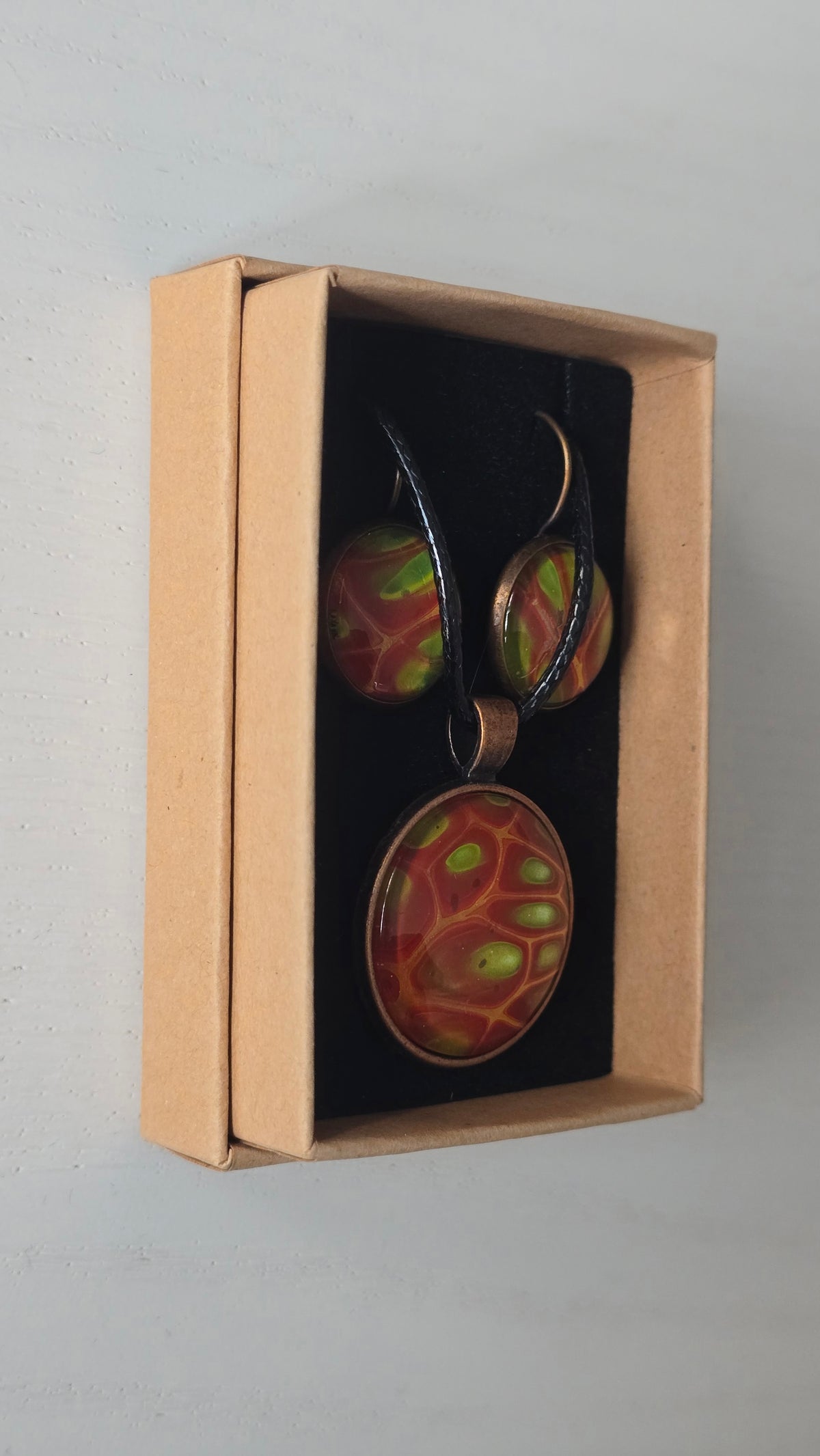 Cabochon-Schmuckset mit rundem Anhänger und zwei Ohrringen mit organischem Muster in Rostrot, Orange und leuchtendem Grün, kupferfarbenen Fassungen und schwarzem Textilband.