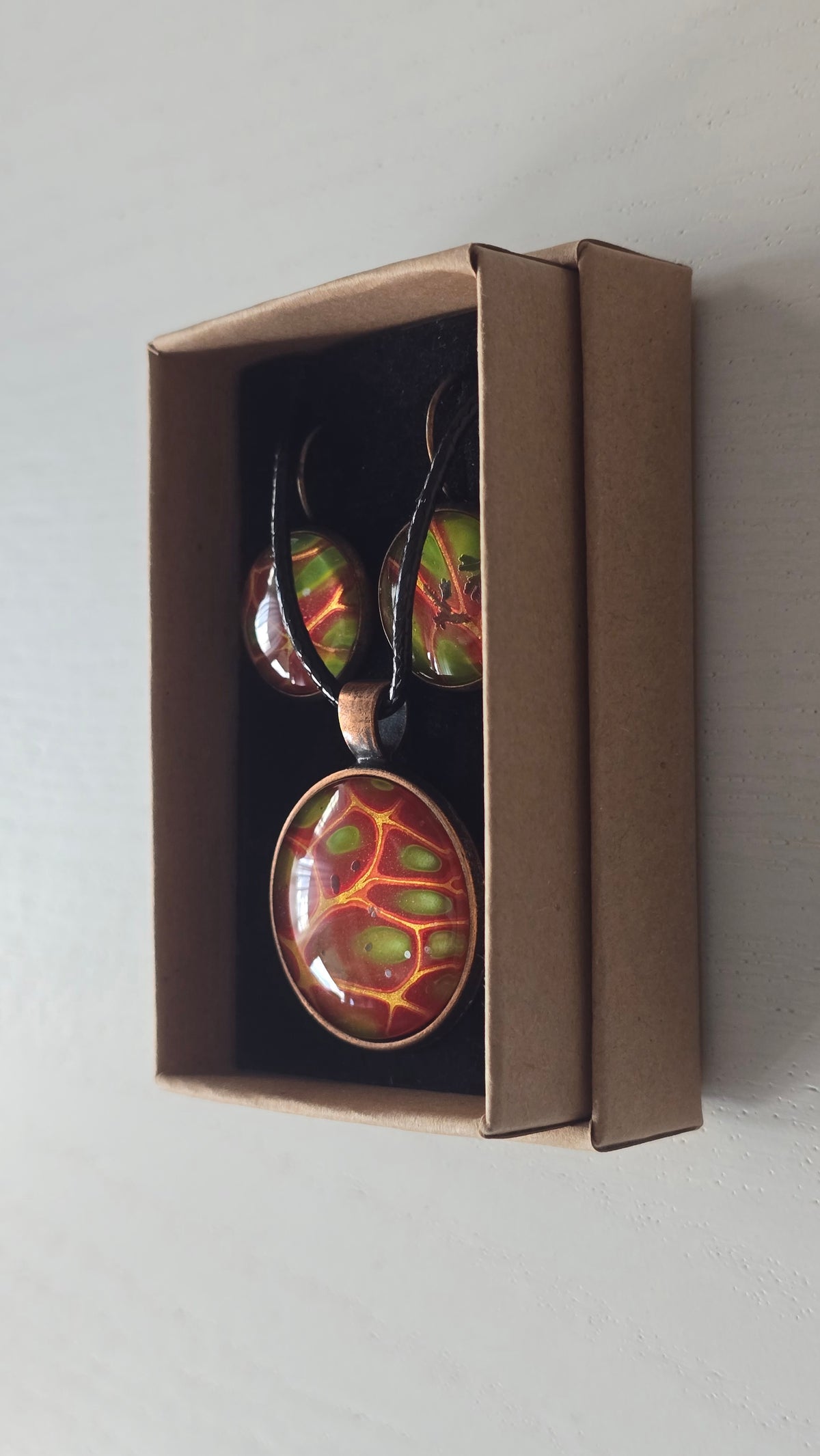 Cabochon-Schmuckset mit rundem Anhänger und zwei Ohrringen mit organischem Muster in Rostrot, Orange und leuchtendem Grün, kupferfarbenen Fassungen und schwarzem Textilband.