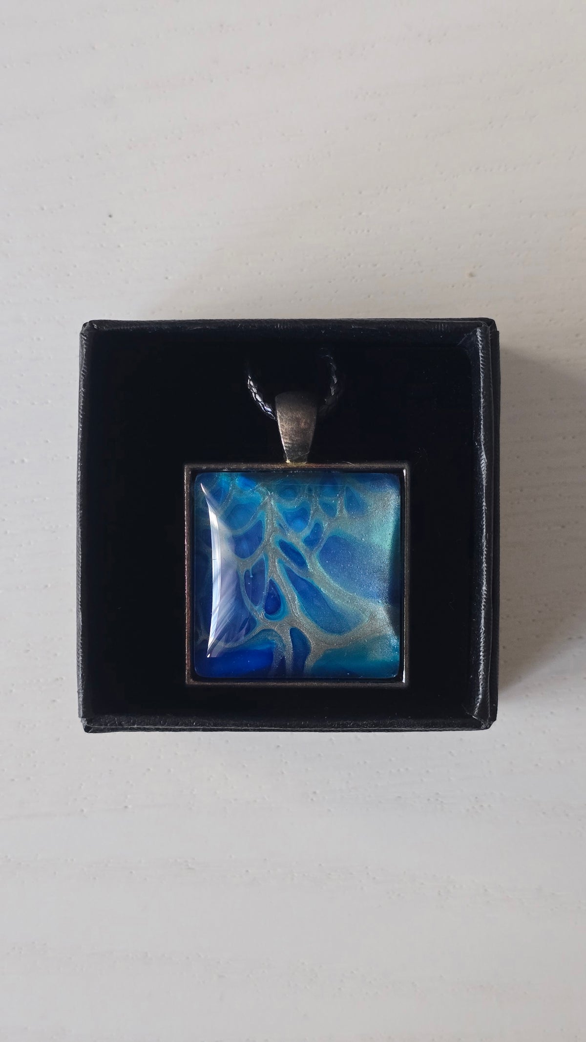 Quadratischer Cabochon-Anhänger an schwarzem Band mit abstraktem, fliessendem Muster in intensiven Blau-, Türkis- und hell schimmernden Nuancen.