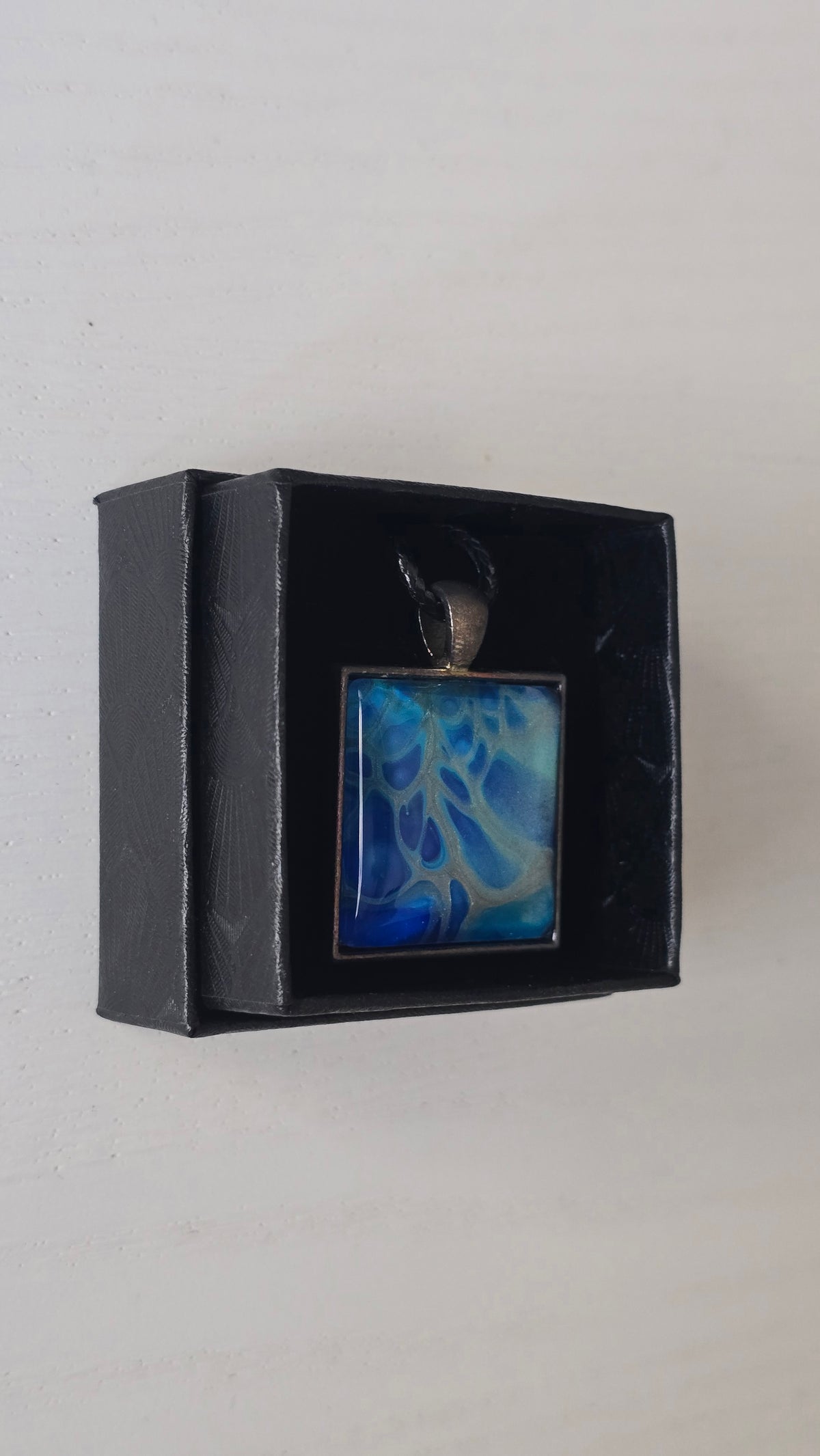 Quadratischer Cabochon-Anhänger an schwarzem Band mit abstraktem, fliessendem Muster in intensiven Blau-, Türkis- und hell schimmernden Nuancen.