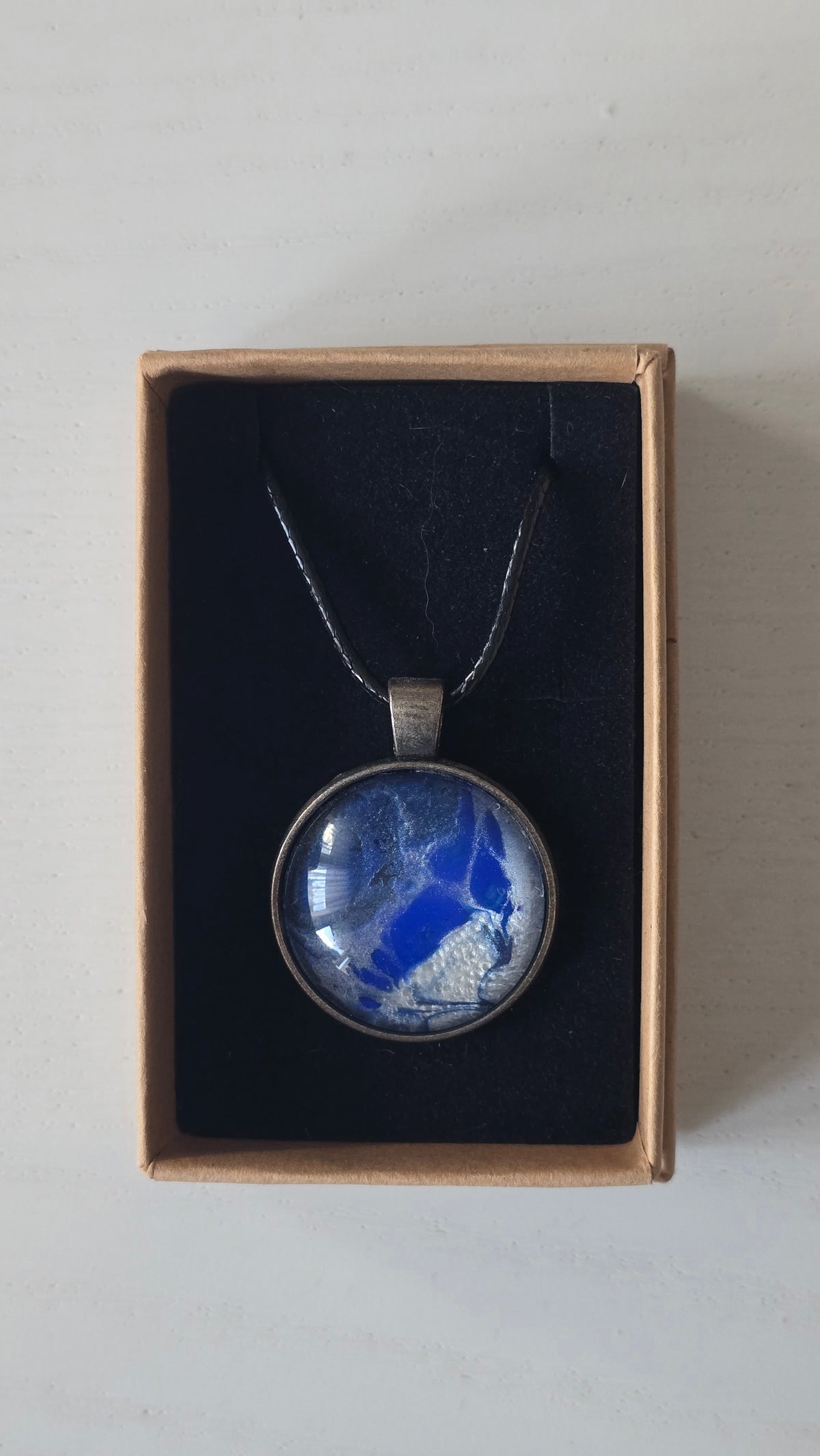 Runde Cabochon Kette Solas Blue Glacier in Kobaltblau, Silber und Weiss mit dunkler Metallfassung und schwarzem Band.