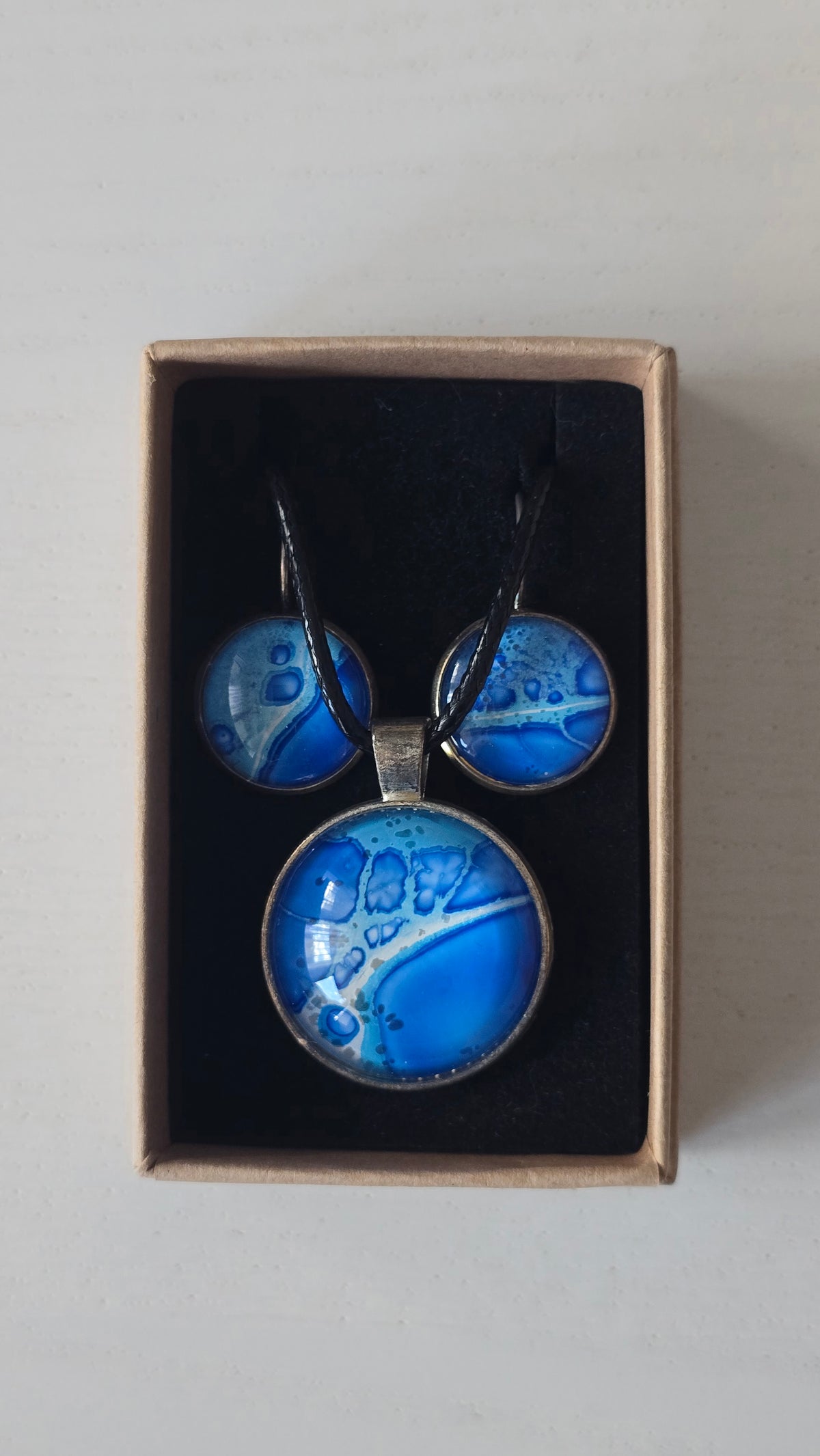 Rundes Cabochon-Schmuckset mit Anhänger und Ohrringen an schwarzem Band, in Kobaltblau, Cyan und Türkis mit runden Zellformen und dunkelblauen Konturen.