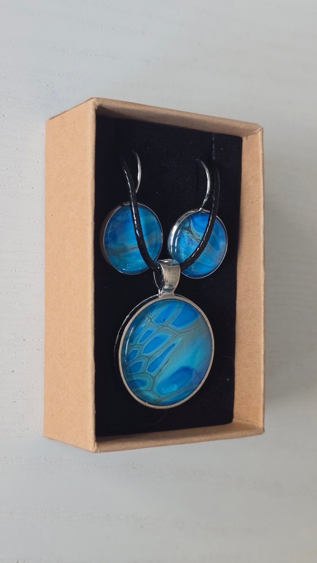Rundes Cabochon-Schmuckset mit Anhänger und Ohrringen an schwarzem Band, mit organischem Muster in Türkis, Cyan und Blau sowie silbernem Schimmer.