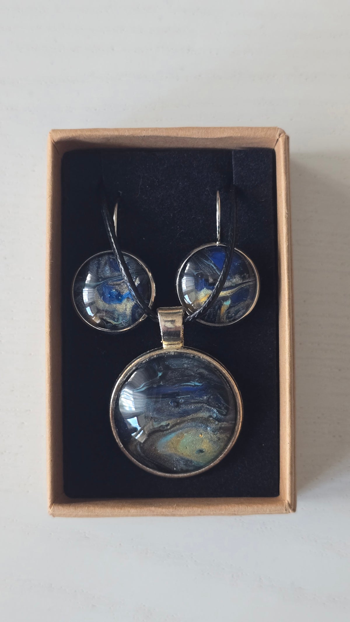 Rundes Cabochon-Schmuckset mit Anhänger und Ohrringen an schwarzem Band, mit fliessendem Muster in Dunkelblau, Schwarz und Petrol sowie goldenen Akzenten.