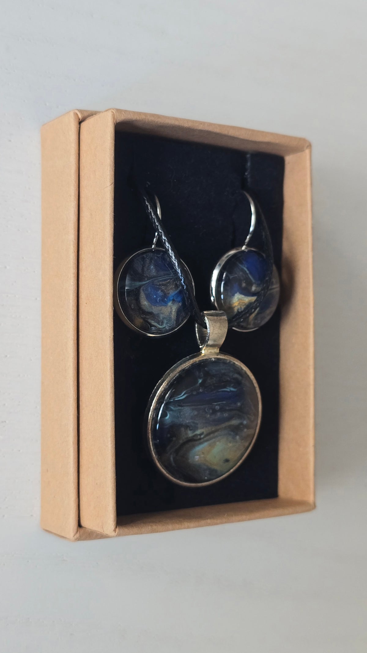 Rundes Cabochon-Schmuckset mit Anhänger und Ohrringen an schwarzem Band, mit fliessendem Muster in Dunkelblau, Schwarz und Petrol sowie goldenen Akzenten.