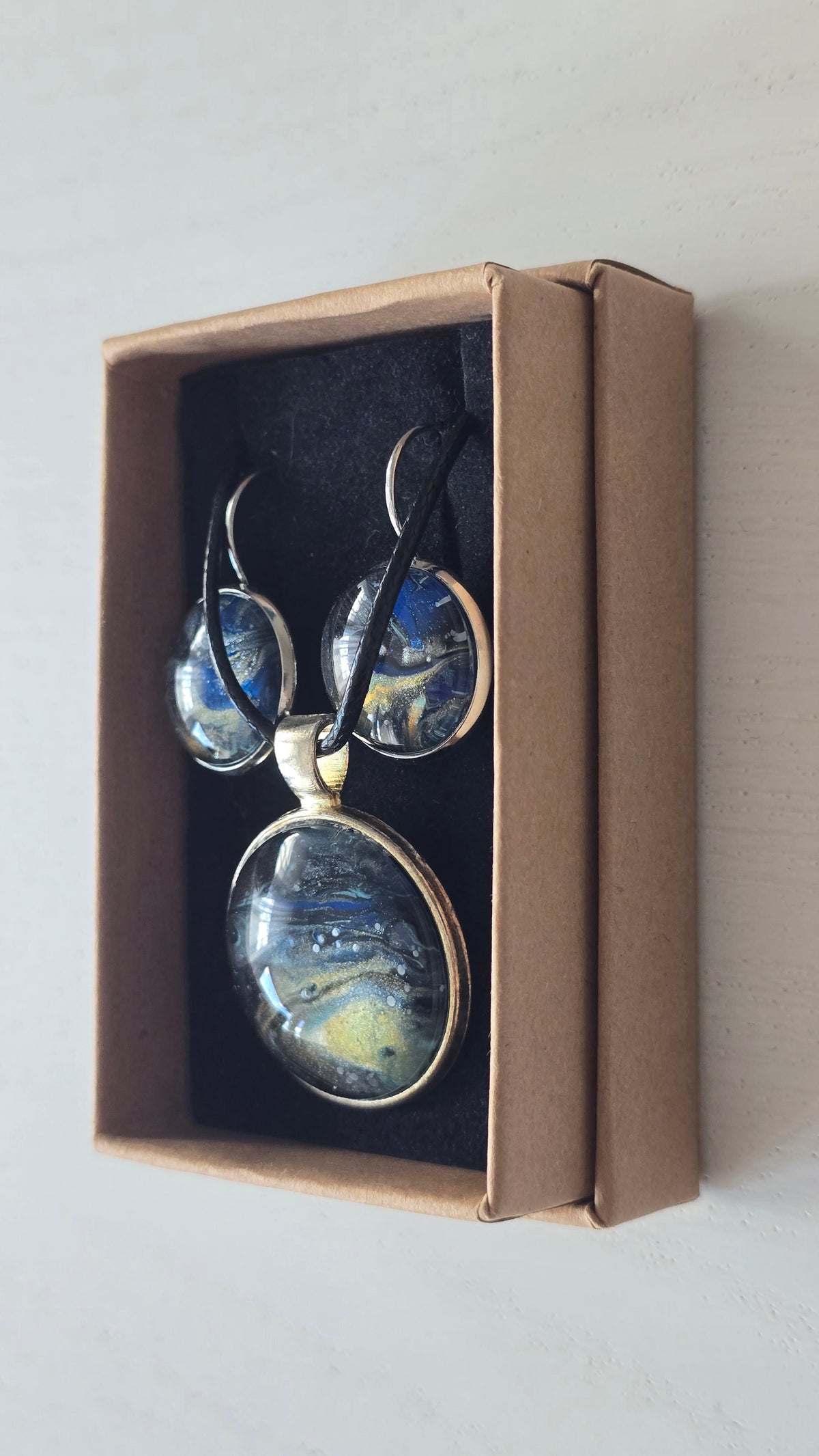 Rundes Cabochon-Schmuckset mit Anhänger und Ohrringen an schwarzem Band, mit fliessendem Muster in Dunkelblau, Schwarz und Petrol sowie goldenen Akzenten.