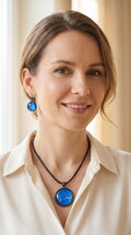 Frau trägt rundes Cabochon-Schmuckset mit Anhänger und Ohrringen in intensivem Blau und Türkis, durchzogen von feinen silbernen Strukturen, an schwarzem geflochtenem Band.