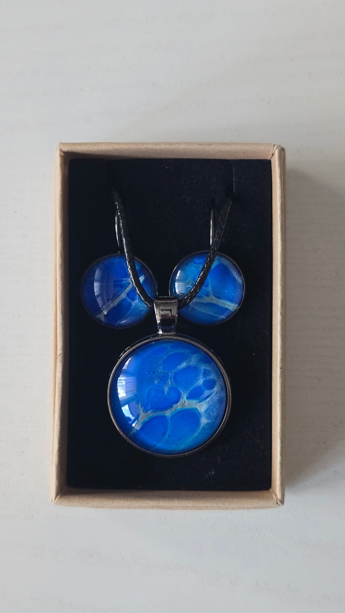 Rundes Cabochon-Schmuckset mit Anhänger und Ohrringen in intensivem Blau und Türkis, durchzogen von feinen silbernen Strukturen, an schwarzem geflochtenem Band.