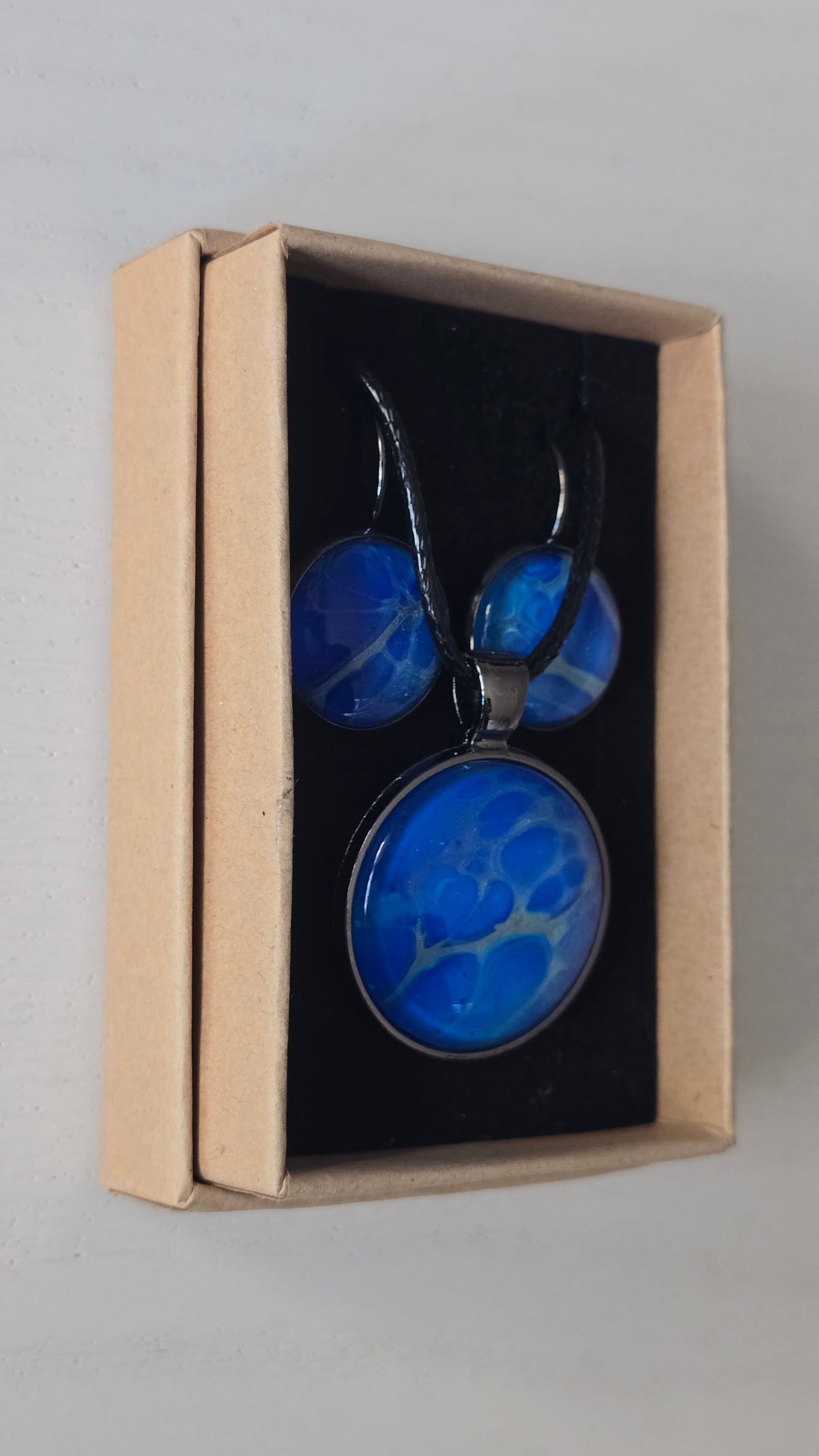 Rundes Cabochon-Schmuckset mit Anhänger und Ohrringen in intensivem Blau und Türkis, durchzogen von feinen silbernen Strukturen, an schwarzem geflochtenem Band.
