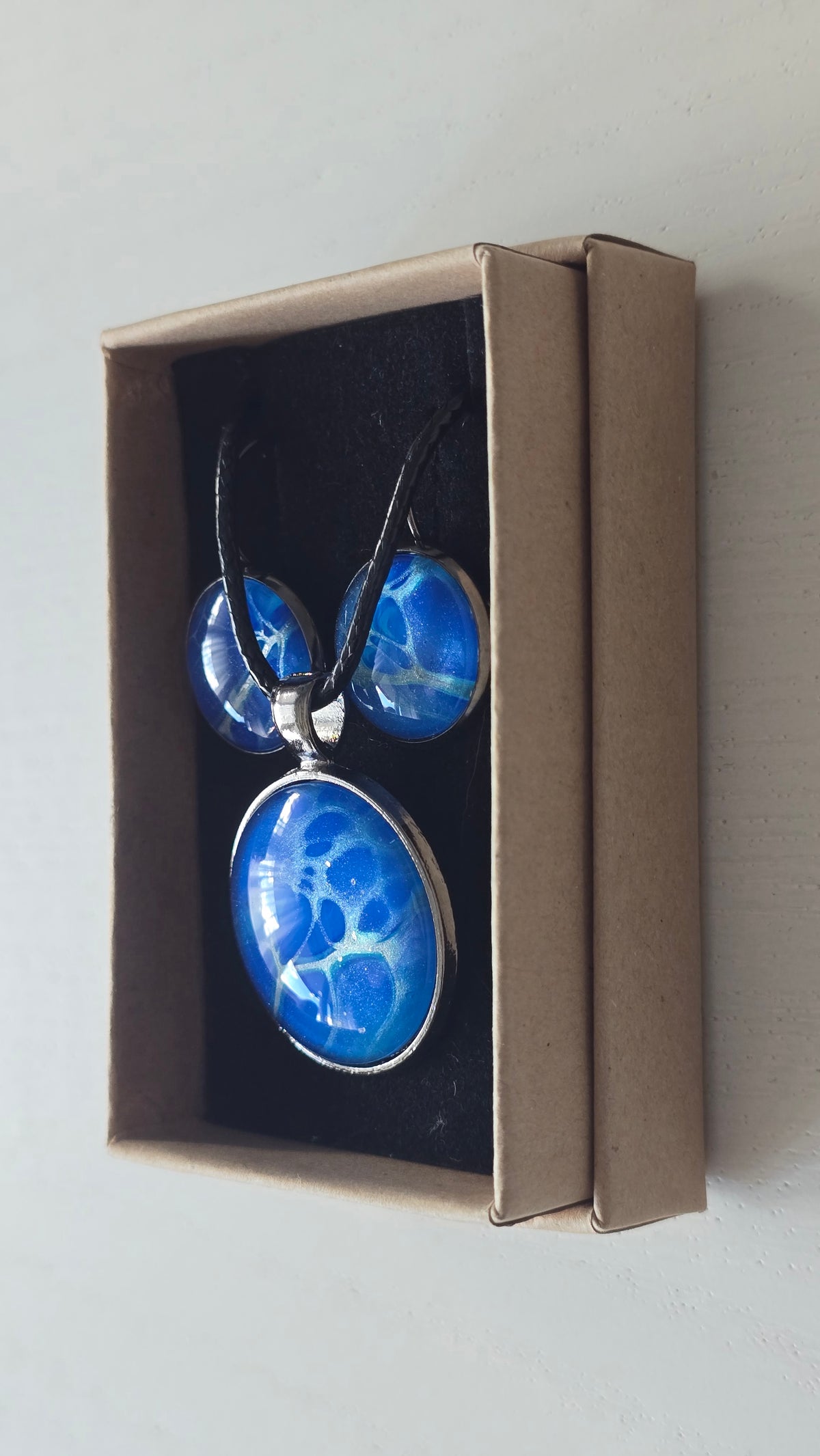 Rundes Cabochon-Schmuckset mit Anhänger und Ohrringen in intensivem Blau und Türkis, durchzogen von feinen silbernen Strukturen, an schwarzem geflochtenem Band.