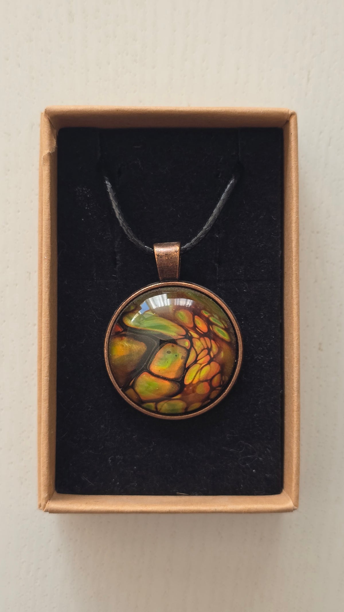 Runde Cabochon Kette Solas Forest Flame in Grün, Orange und Gold mit dunklen Strukturen, bronzefarbener Fassung und schwarzem Band.