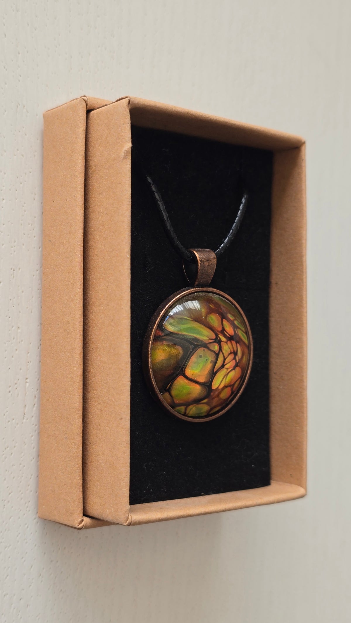 Runde Cabochon Kette Solas Forest Flame in Grün, Orange und Gold mit dunklen Strukturen, bronzefarbener Fassung und schwarzem Band.