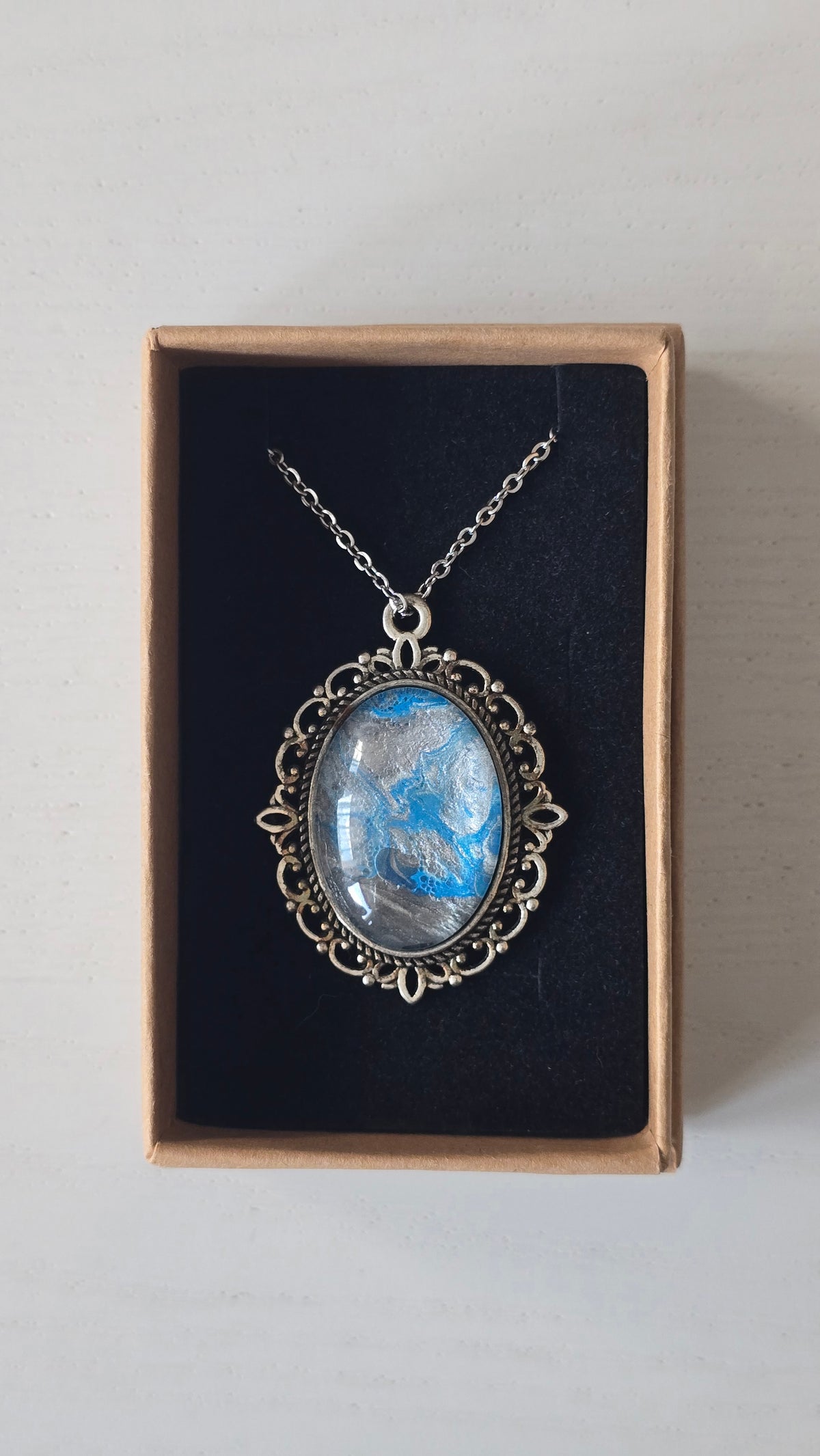 Ovale Cabochon Kette Solas Frozen Current in leuchtendem Blau, Silber und Grau mit verzierter silberfarbener Fassung und Metallkette.
