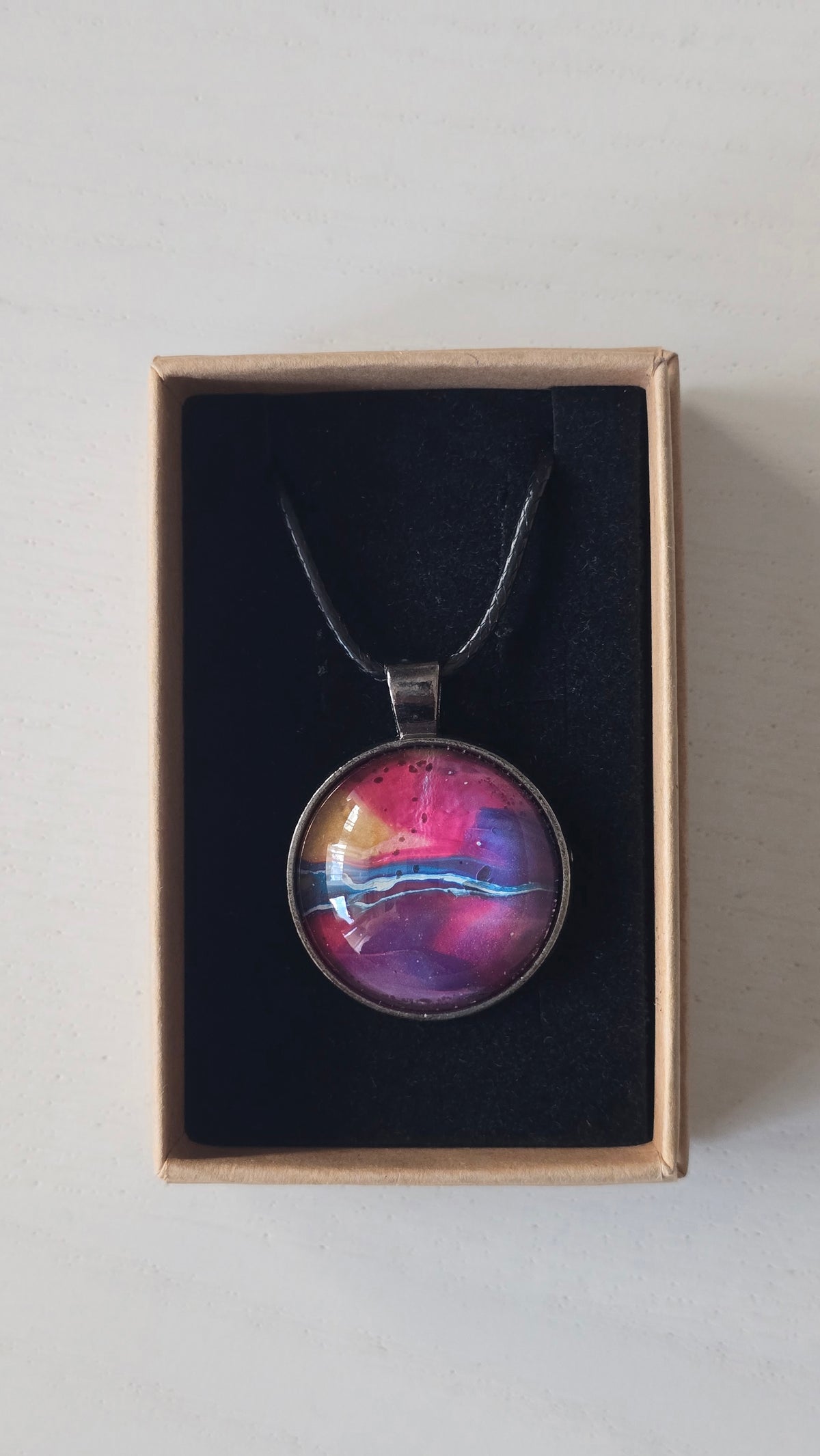 Runder Cabochon-Anhänger an schwarzem Band mit geschichtetem Muster in Fuchsia, Pink, Violett, Blau, Türkis und einem goldenen Akzent.