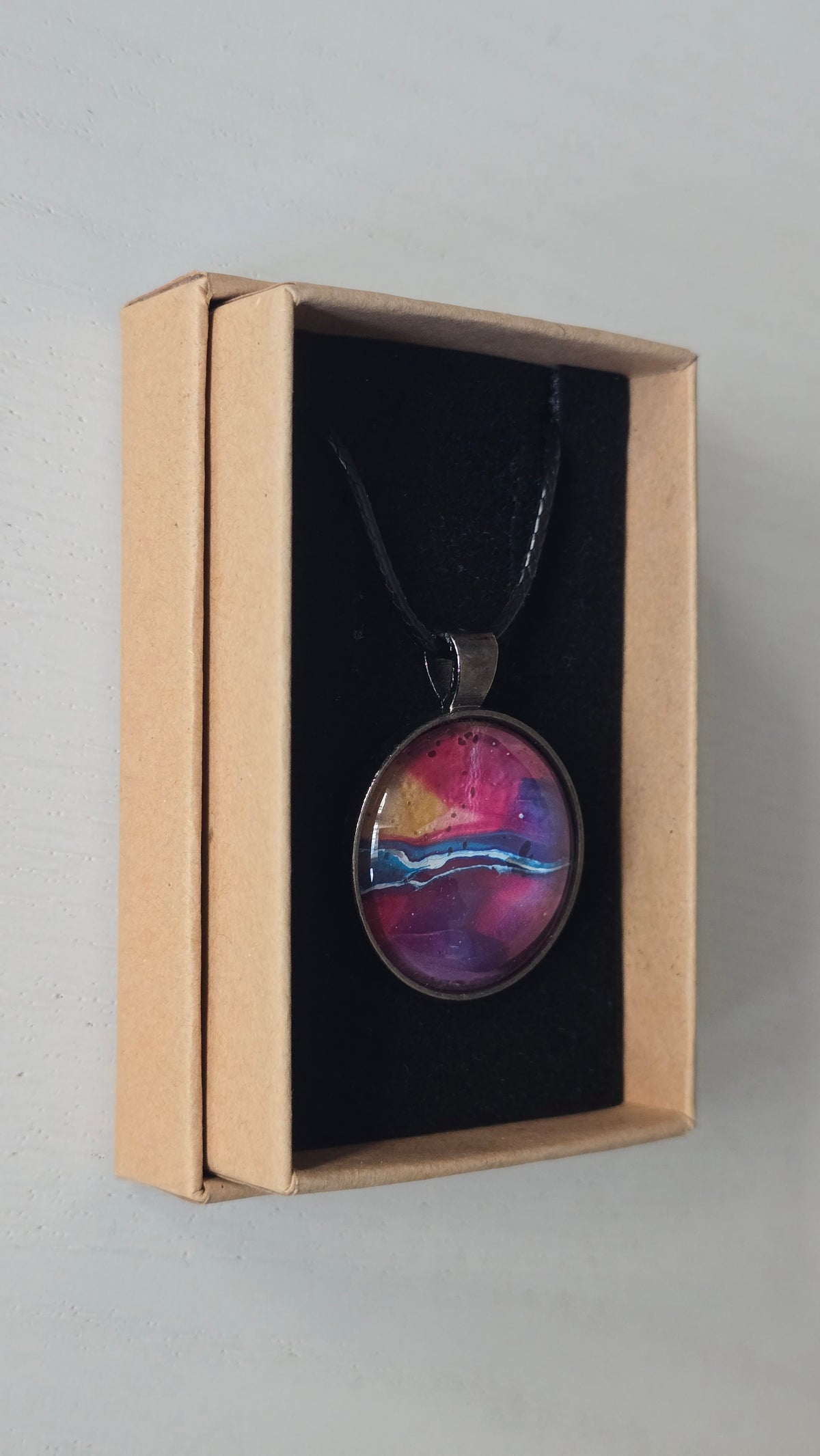Runder Cabochon-Anhänger an schwarzem Band mit geschichtetem Muster in Fuchsia, Pink, Violett, Blau, Türkis und einem goldenen Akzent.