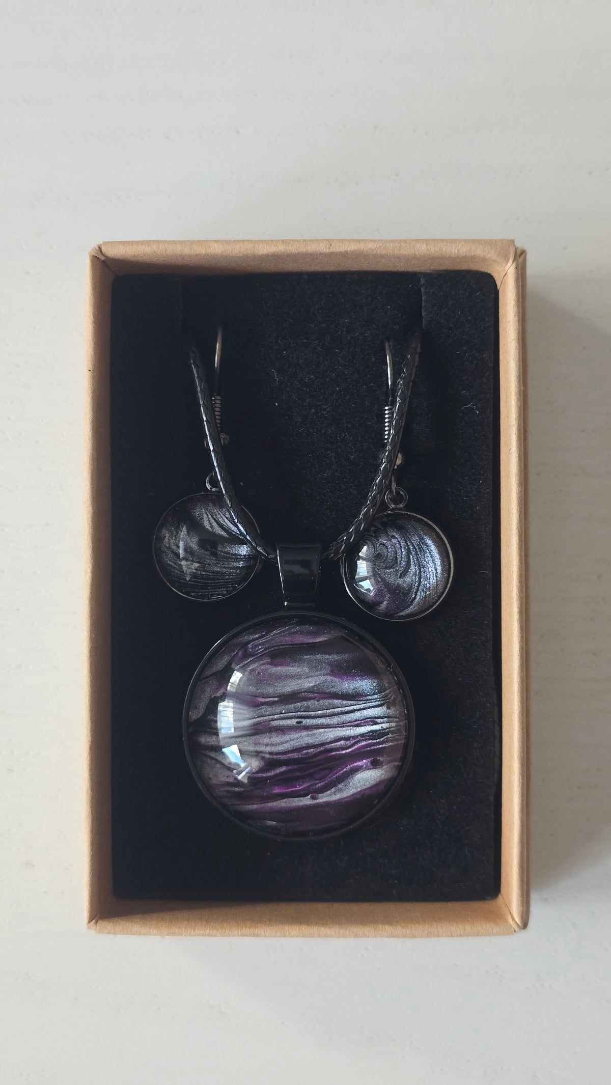 Rundes Cabochon-Schmuckset mit Anhänger und Ohrringen an schwarzem Band, mit geschichtetem Muster in Schwarz, Anthrazit, Violett und silbrigem Schimmer.