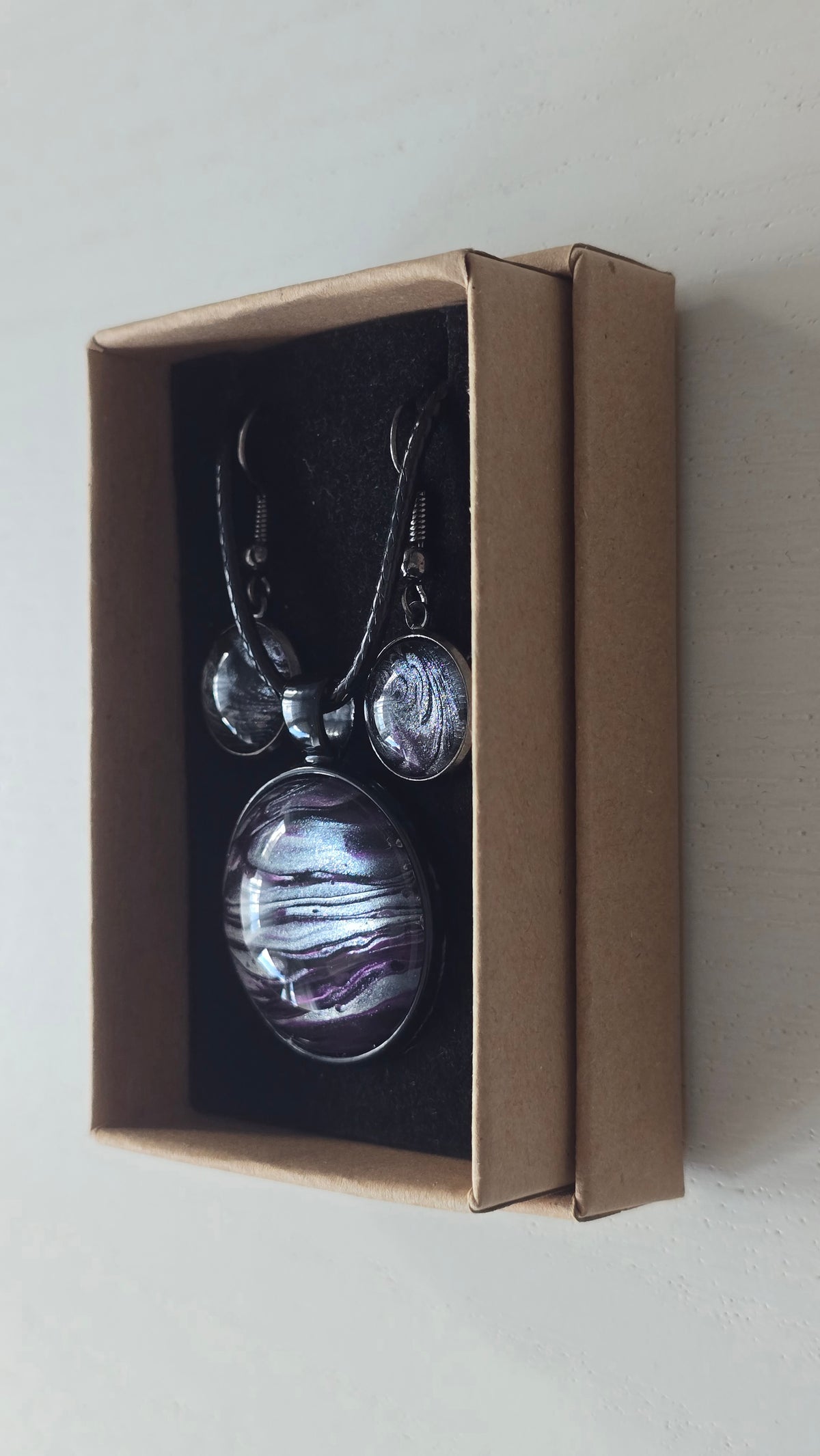 Rundes Cabochon-Schmuckset mit Anhänger und Ohrringen an schwarzem Band, mit geschichtetem Muster in Schwarz, Anthrazit, Violett und silbrigem Schimmer.