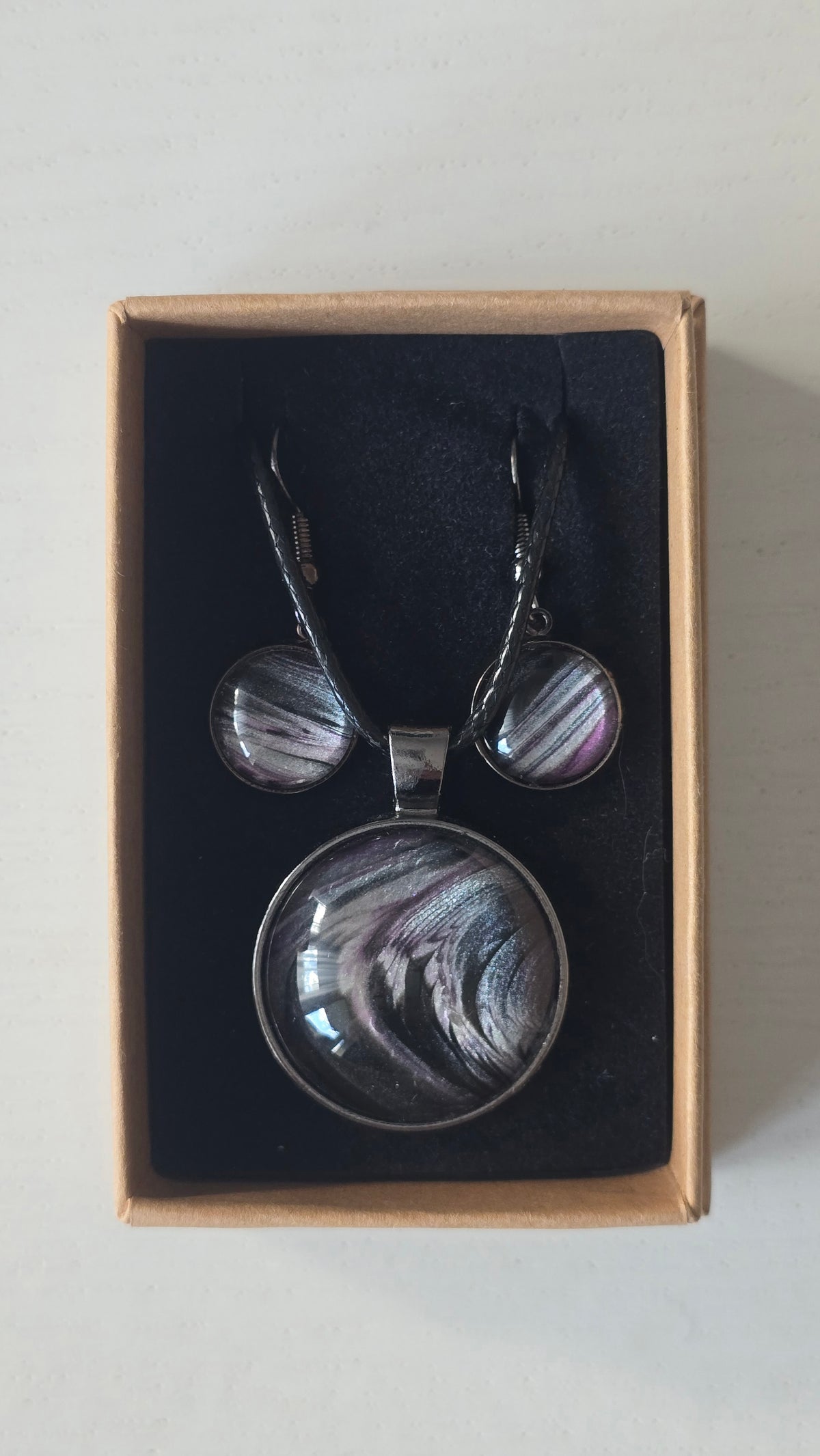 Cabochon-Schmuckset mit rundem Anhänger und zwei Ohrringen in Schwarz und Anthrazit mit geschwungenem silbernem Muster und violetten Akzenten.
