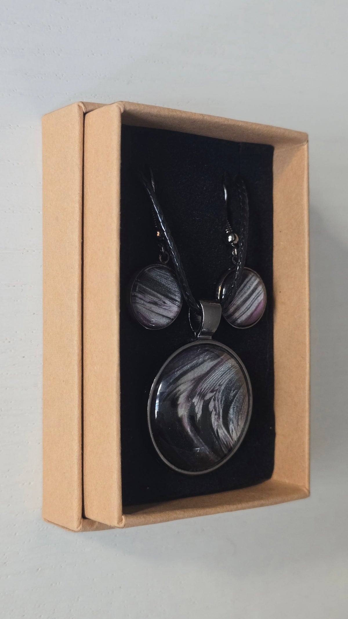 Cabochon-Schmuckset mit rundem Anhänger und zwei Ohrringen in Schwarz und Anthrazit mit geschwungenem silbernem Muster und violetten Akzenten.