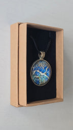 Runder Cabochon-Anhänger an schwarzem Band mit abstraktem, mineralischem Muster in Blau, Türkis, Weiss und Gold.