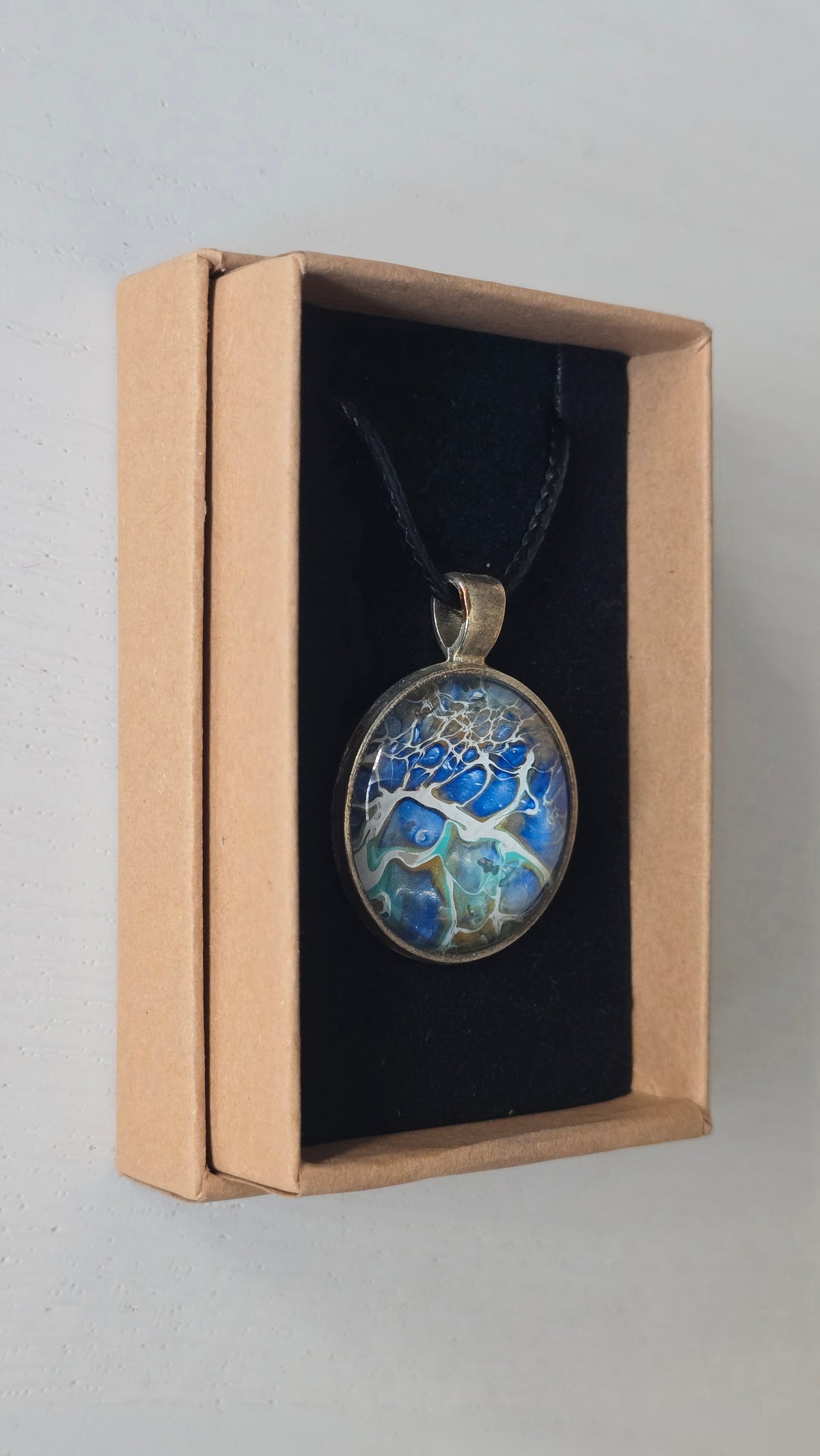 Runder Cabochon-Anhänger an schwarzem Band mit abstraktem, mineralischem Muster in Blau, Türkis, Weiss und Gold.