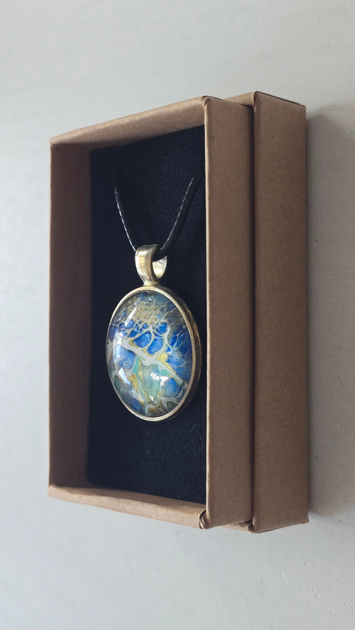 Runder Cabochon-Anhänger an schwarzem Band mit abstraktem, mineralischem Muster in Blau, Türkis, Weiss und Gold.