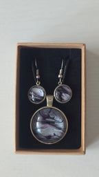 Rundes Cabochon-Schmuckset mit Anhänger und Ohrringen an schwarzem Band, mit silbrigem Perlmuttschimmer, Violett und fliessenden schwarzen Linien.