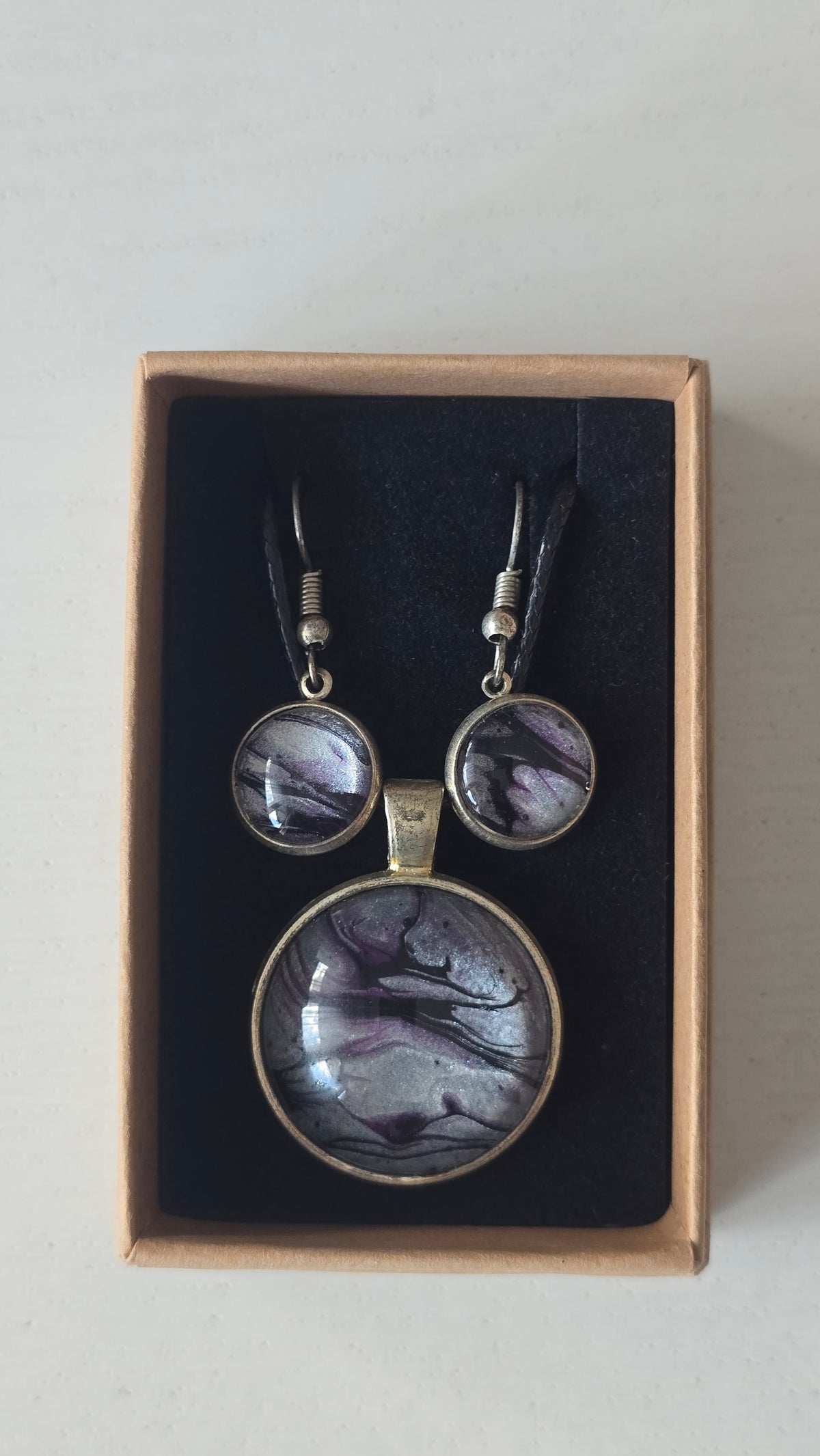 Rundes Cabochon-Schmuckset mit Anhänger und Ohrringen an schwarzem Band, mit silbrigem Perlmuttschimmer, Violett und fliessenden schwarzen Linien.