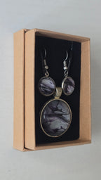 Rundes Cabochon-Schmuckset mit Anhänger und Ohrringen an schwarzem Band, mit silbrigem Perlmuttschimmer, Violett und fliessenden schwarzen Linien.