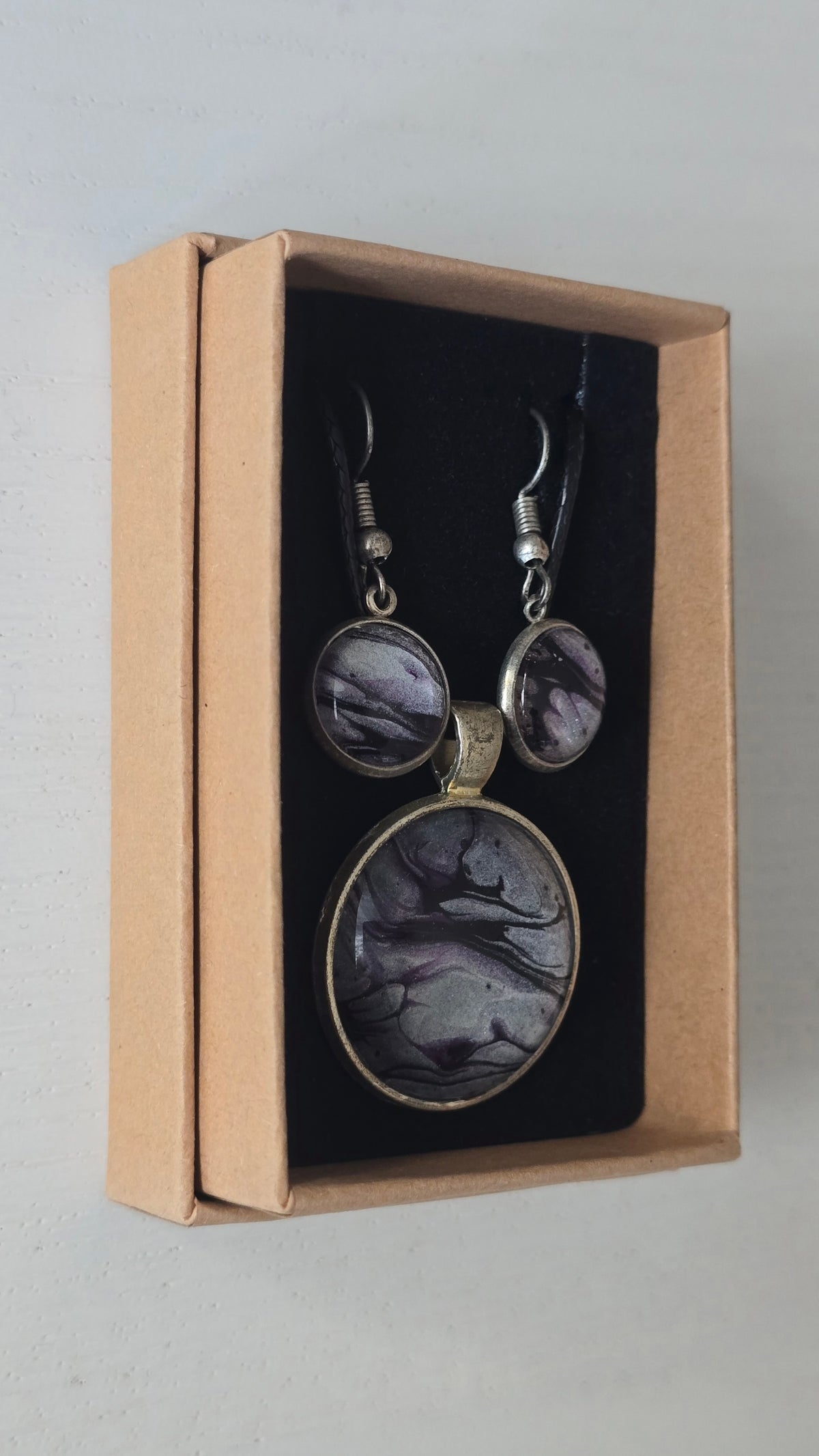 Rundes Cabochon-Schmuckset mit Anhänger und Ohrringen an schwarzem Band, mit silbrigem Perlmuttschimmer, Violett und fliessenden schwarzen Linien.