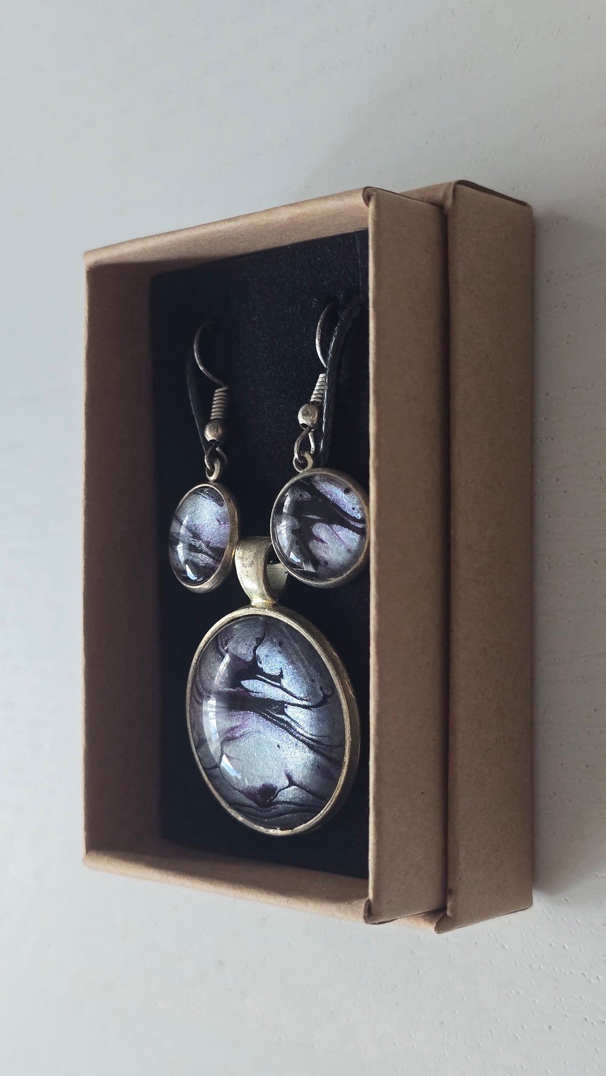 Rundes Cabochon-Schmuckset mit Anhänger und Ohrringen an schwarzem Band, mit silbrigem Perlmuttschimmer, Violett und fliessenden schwarzen Linien.
