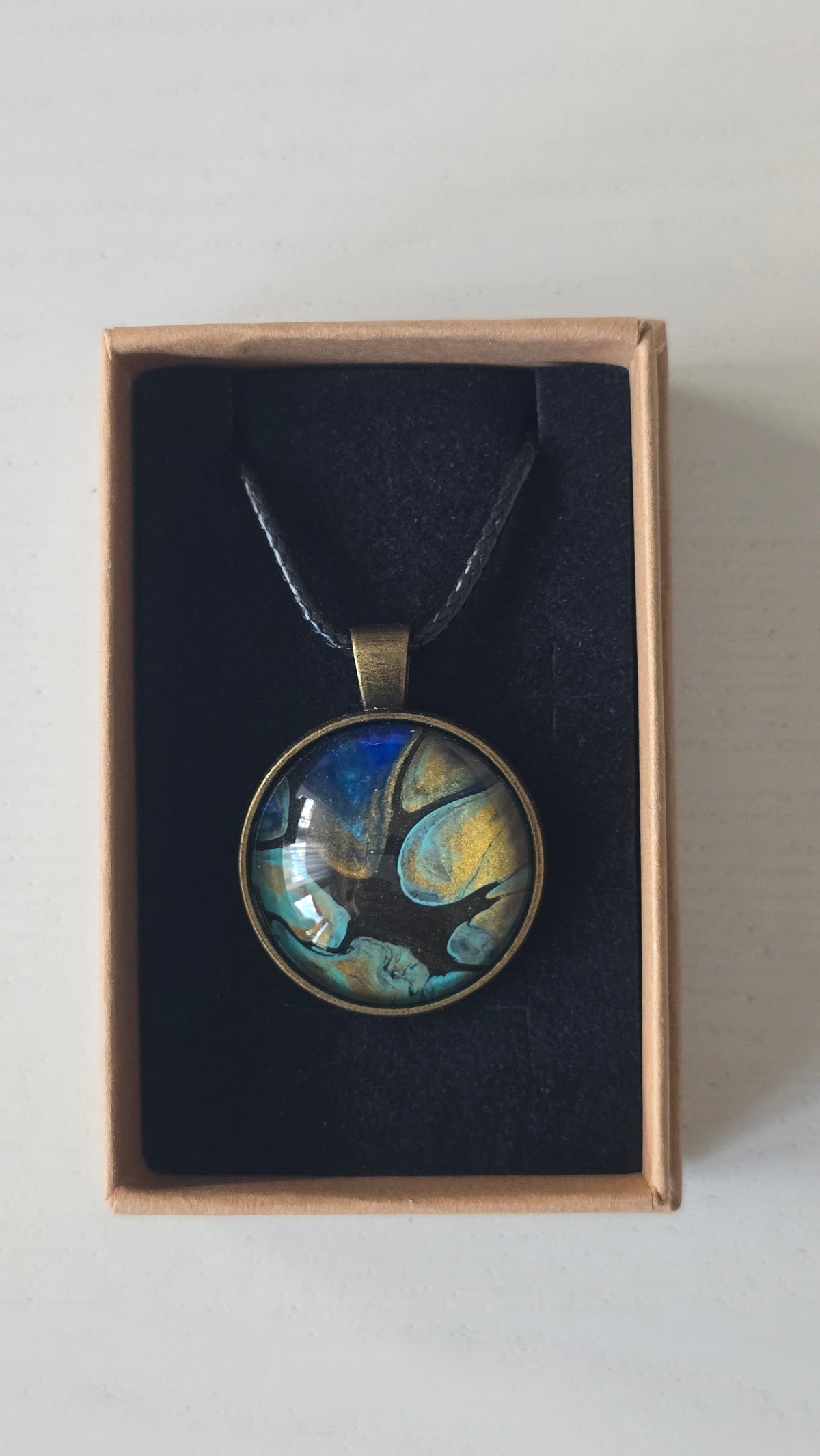 Runder Cabochon-Anhänger an schwarzem Band mit organischem Muster in Türkis, tiefem Blau, Schwarz und warm schimmernden Goldtönen.