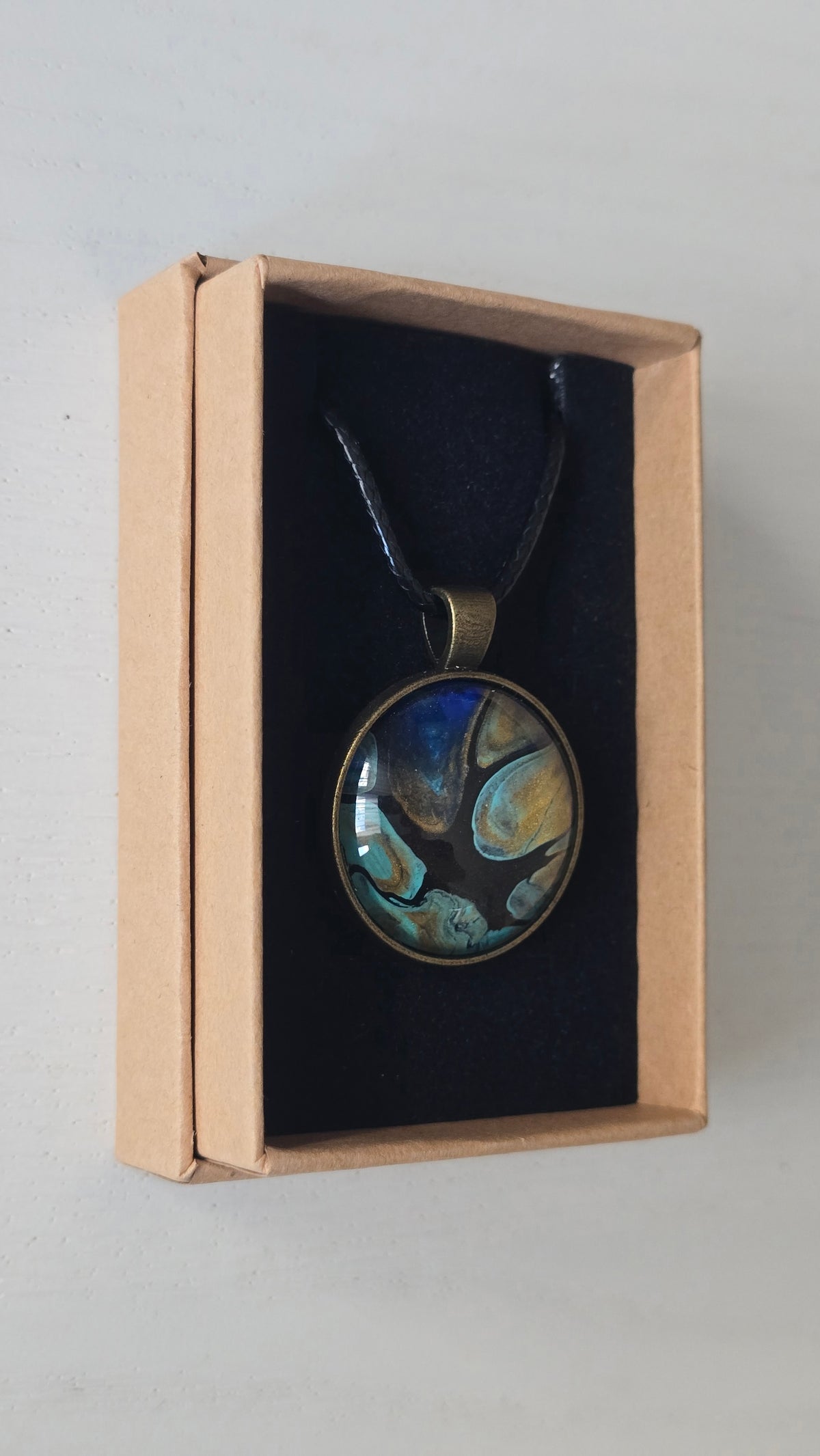 Runder Cabochon-Anhänger an schwarzem Band mit organischem Muster in Türkis, tiefem Blau, Schwarz und warm schimmernden Goldtönen.