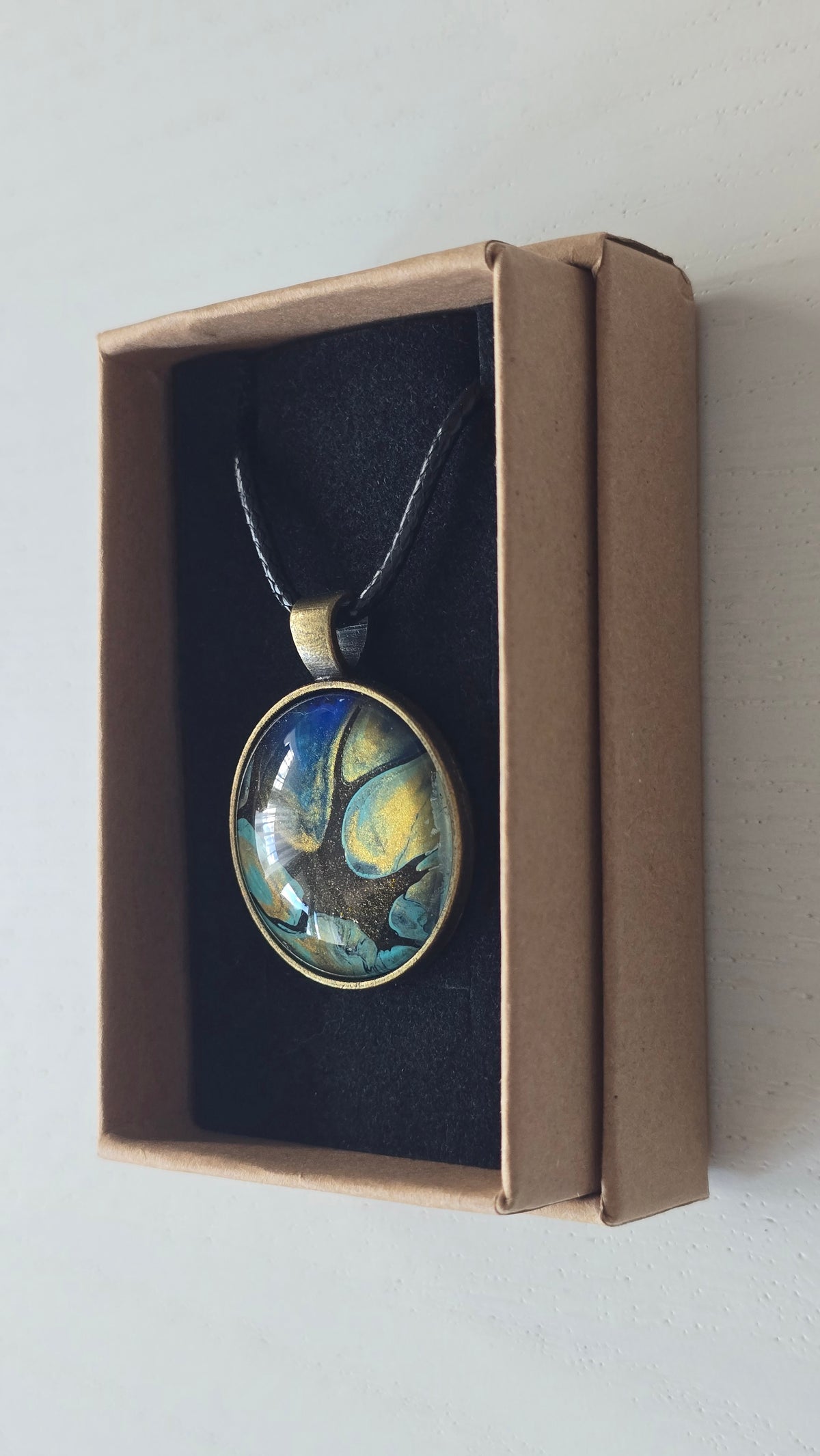 Runder Cabochon-Anhänger an schwarzem Band mit organischem Muster in Türkis, tiefem Blau, Schwarz und warm schimmernden Goldtönen.
