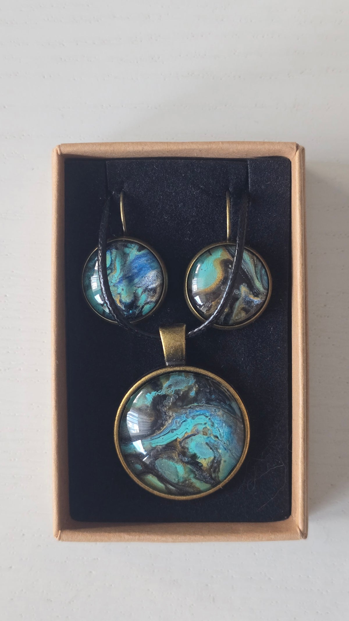 Cabochon-Schmuckset mit rundem Anhänger und zwei Ohrringen mit marmoriertem Muster in Türkis, Petrol, Blau und Schwarz mit goldig-bronzefarbenen Akzenten.