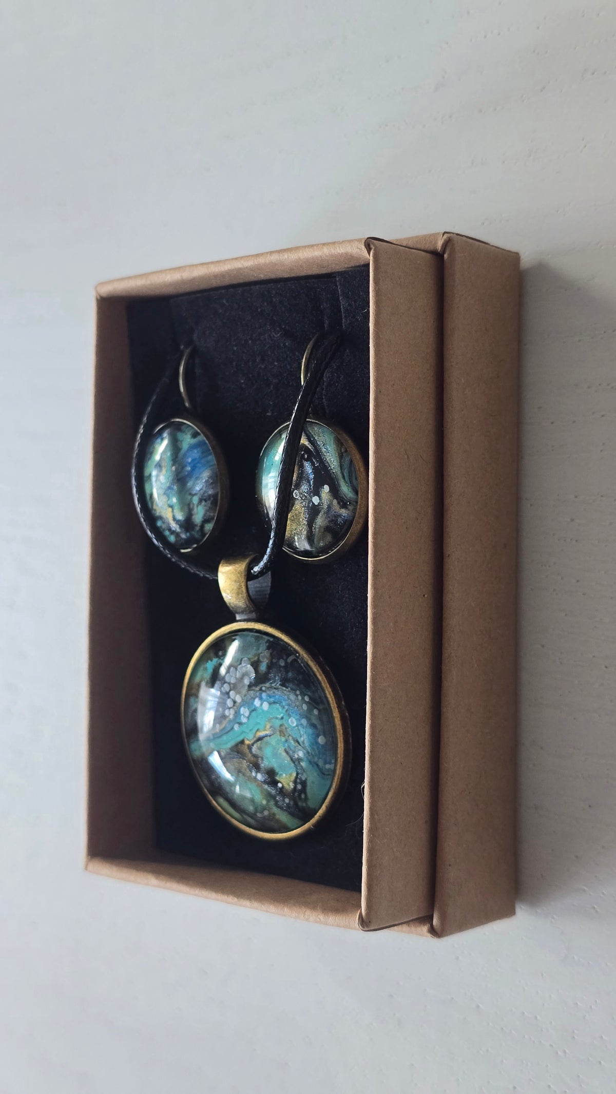 Cabochon-Schmuckset mit rundem Anhänger und zwei Ohrringen mit marmoriertem Muster in Türkis, Petrol, Blau und Schwarz mit goldig-bronzefarbenen Akzenten.