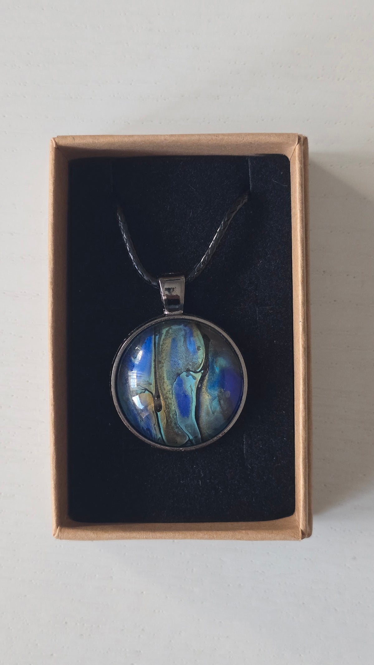 Runde Cabochon Kette Solas Ocean Veil in Blau, Türkis, Petrol und Grün mit dunkler Metallfassung und schwarzem Band.